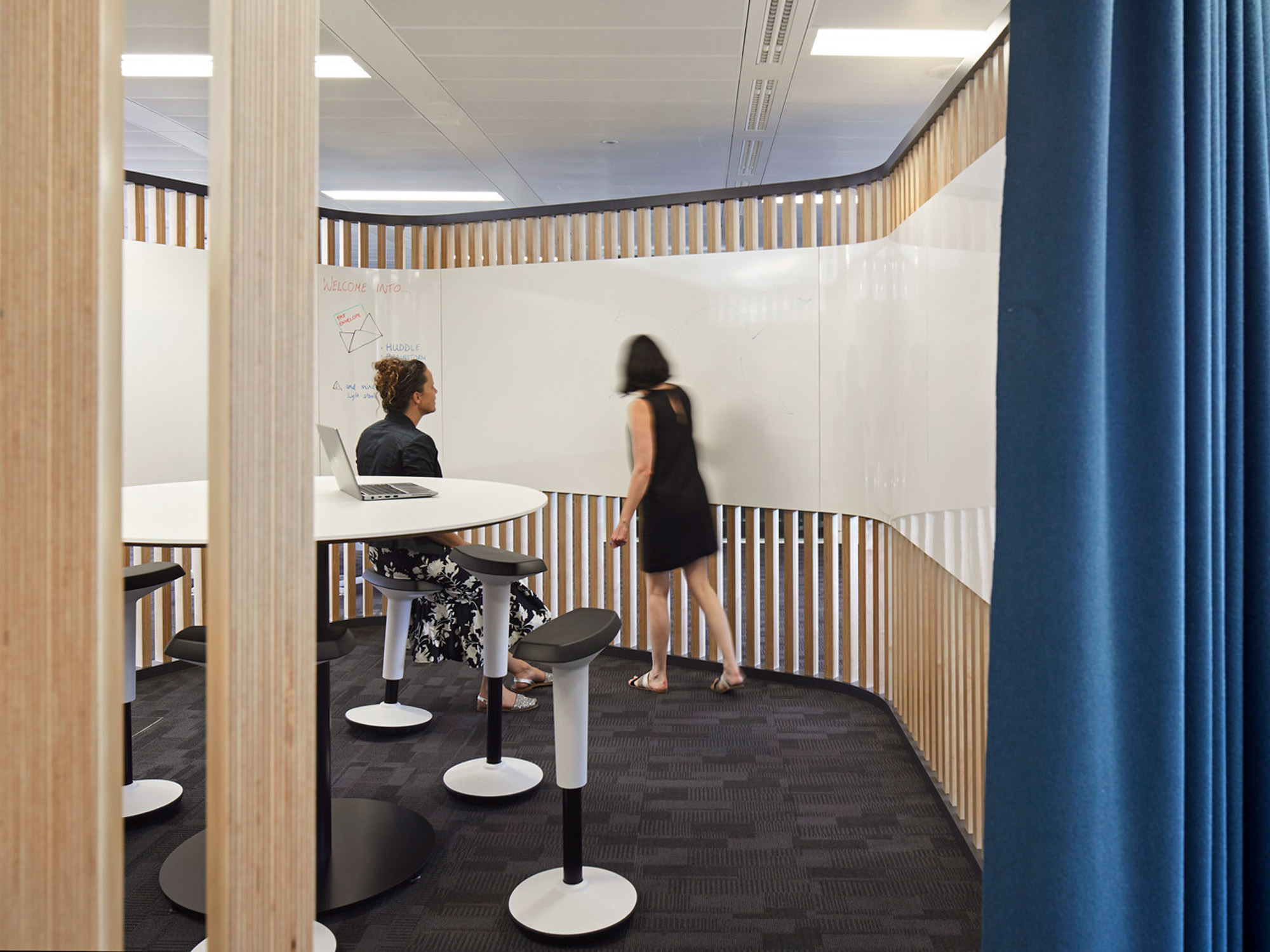 Modern office break area with minimalist white tables and circular black stools. Natural light filters through wooden slatted partitions, enhancing the open yet private ambiance. A whiteboard is mounted on the wall for collaboration.