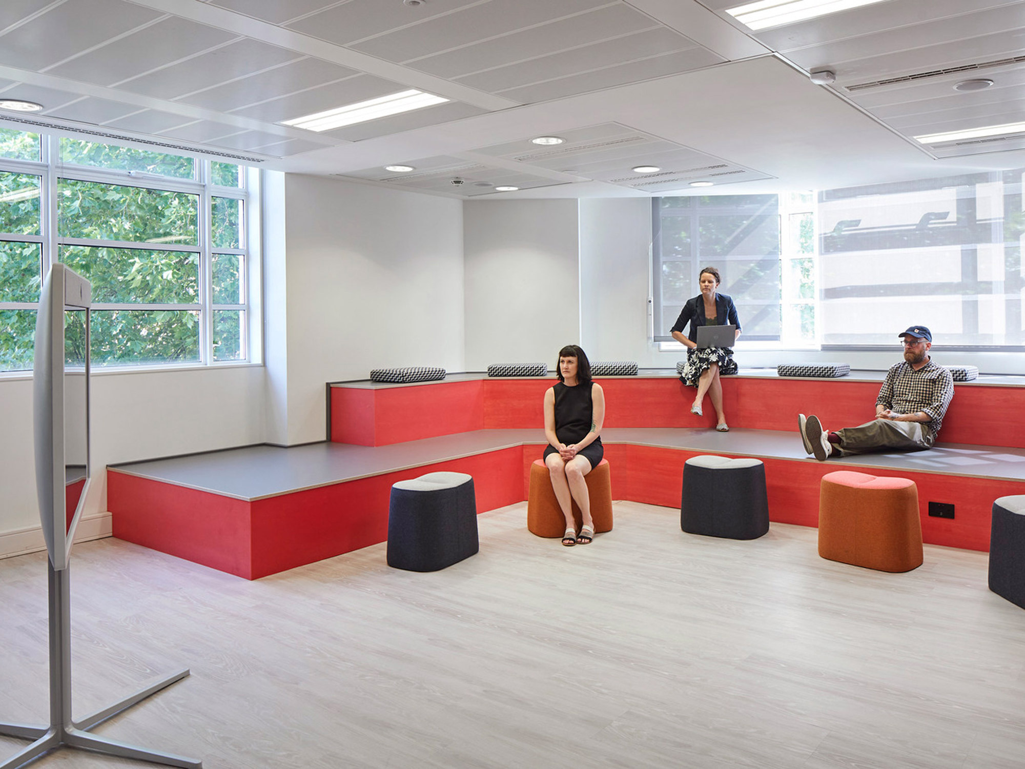 Modern open-concept workspace featuring tiered seating in bold red, gray cushioned accents, and movable brown poufs. Natural light filters through floor-to-ceiling windows complementing the minimalist decor and enhancing the versatility of the multipurpose area.