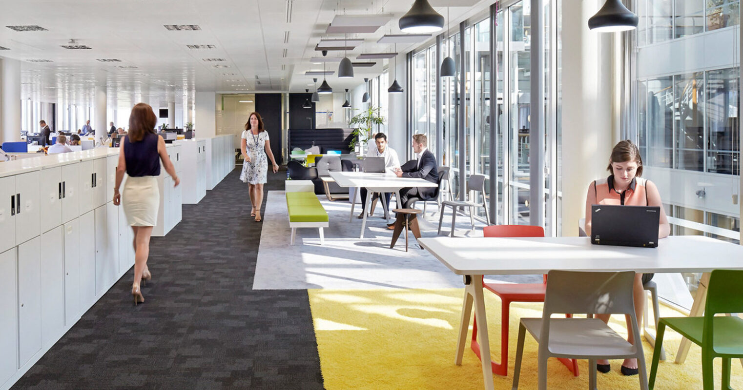 Sunlight streams into a modern office space featuring long white desks, colorful ergonomic chairs, and pendant lighting. Employees work at spaced-out stations promoting productivity and social distancing. The vibrant yellow carpet adds a cheerful accent to the clean, minimalist design.