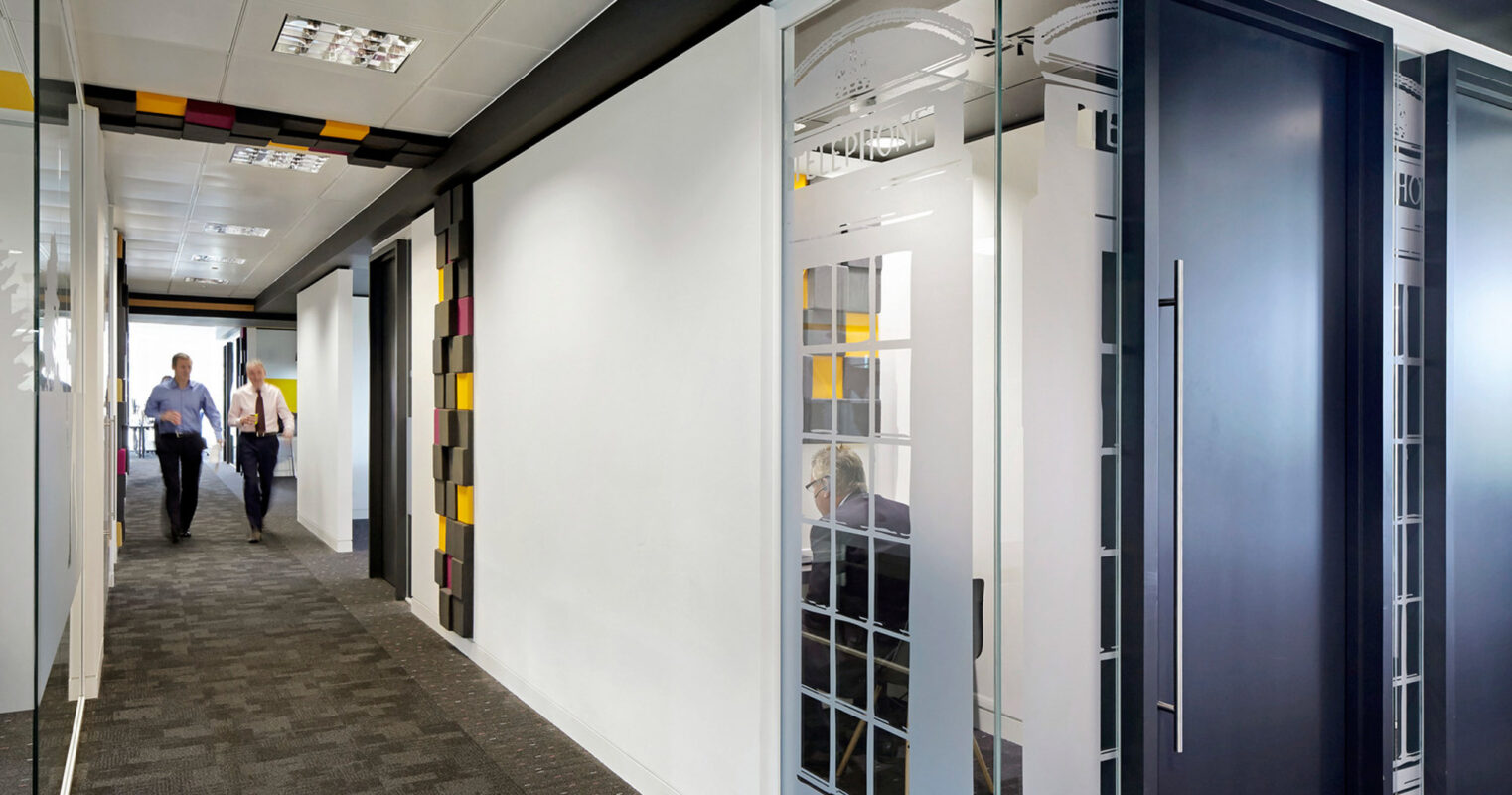 Modern office interior featuring a neutral color palette with splashes of color from rectangular wall panels. Glass partitions with metallic frames provide transparency. Slat ceilings and recessed lighting accentuate the elongated hallway where office workers move about.
