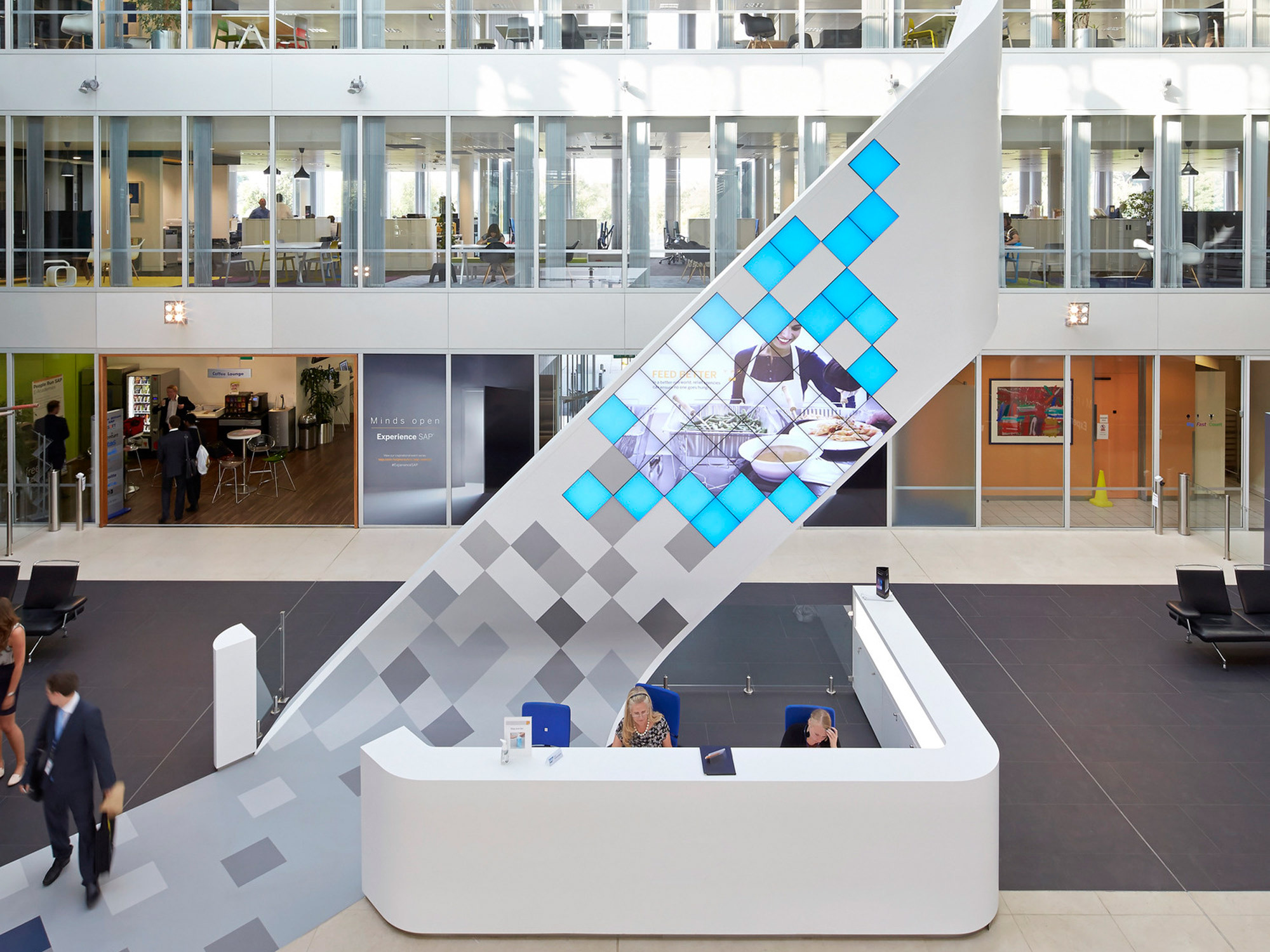 Modern office lobby featuring a sculptural white staircase with blue-tinted glass balustrades. The reception area below mirrors the angular design, complemented by a patterned gray floor that extends throughout the bright atrium surrounded by multi-level glass-fronted offices.