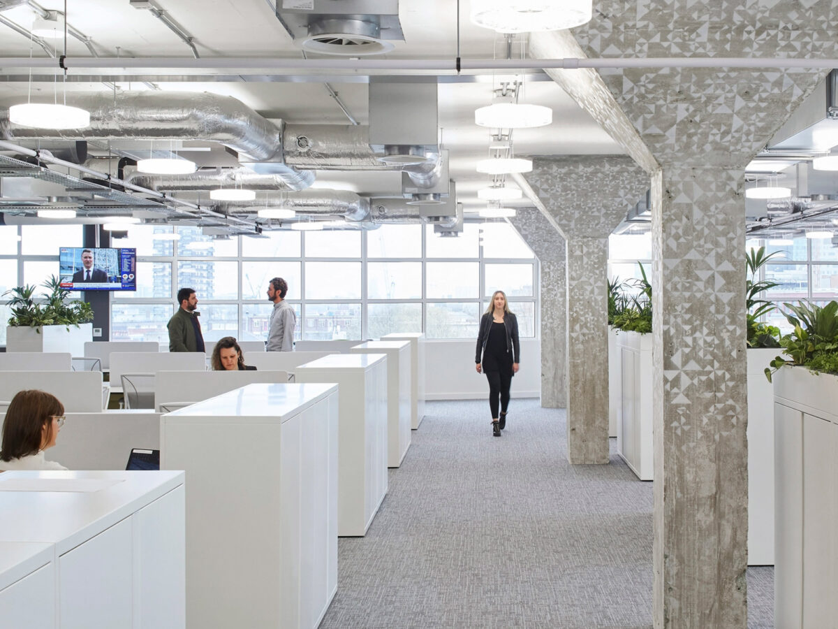 Open-plan office space featuring white minimalist desks, ergonomic chairs, and privacy panels. Exposed concrete columns and ceiling add an industrial chic vibe, while large windows provide ample natural light. Employees are casually dressed, indicating a contemporary, collaborative work environment.