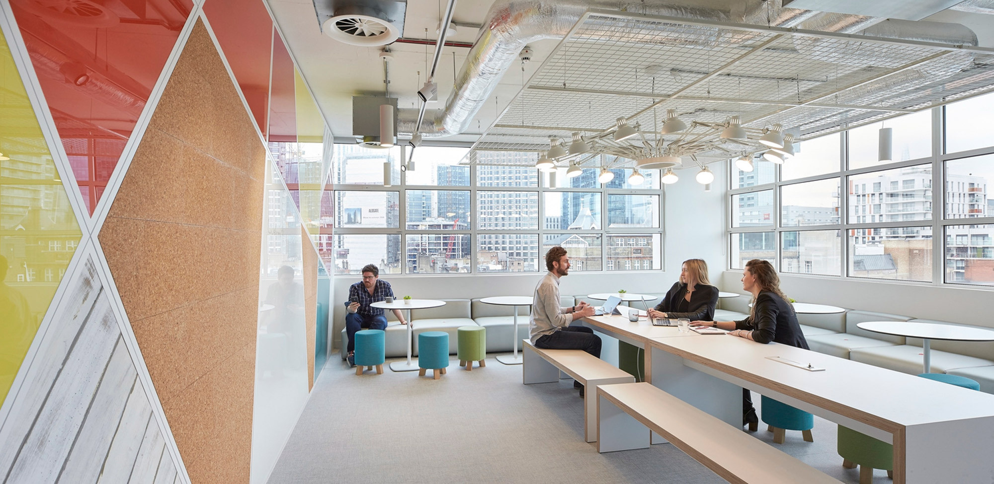 Modern office space boasting an open floor plan with exposed ceiling ductwork, incorporating natural light through large windows. Vibrant color blocks on glass partitions blend with neutral tones, while varied seating options foster collaborative and individual workstations.