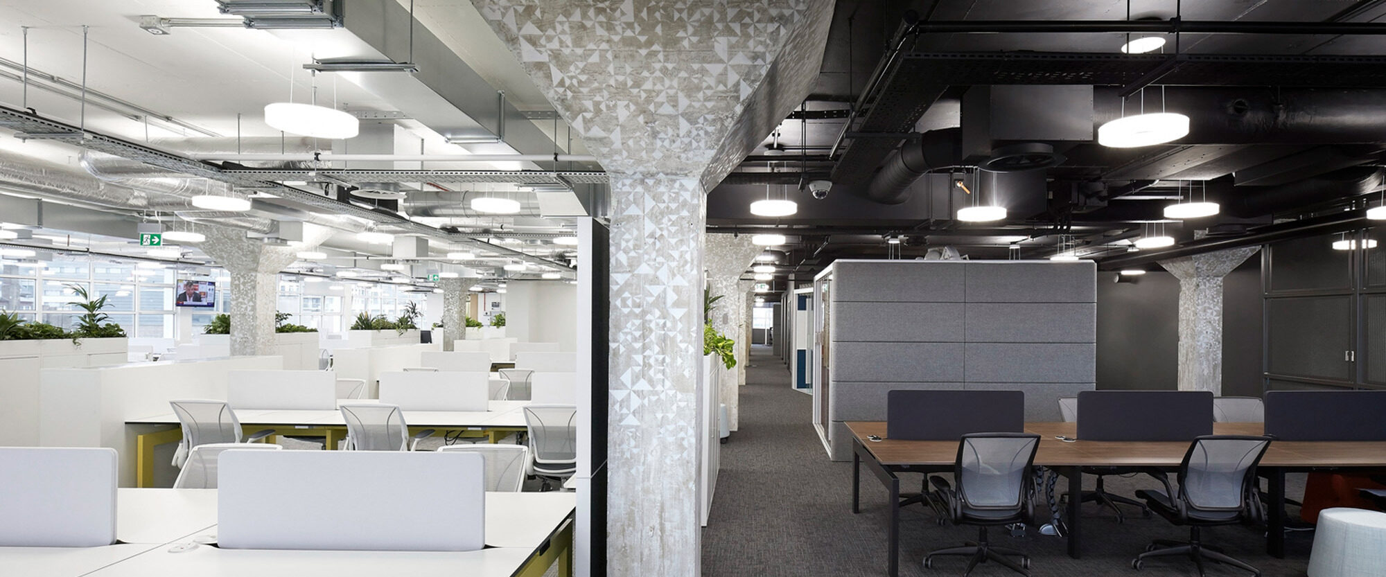 Modern office interior featuring an open-plan layout with white desks, ergonomic chairs, and partition panels for privacy. Exposed ceiling ductwork contrasts with decorative white lattice columns, while pendant lights illuminate the space evenly.