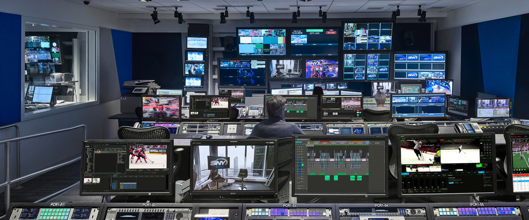 Modern broadcast control room featuring rows of advanced video monitoring equipment, multiple screens displaying various channels, and an operator focused on adjusting technical settings. The space is organized, reflecting a high-tech and efficient design with ergonomic seating and task lighting.