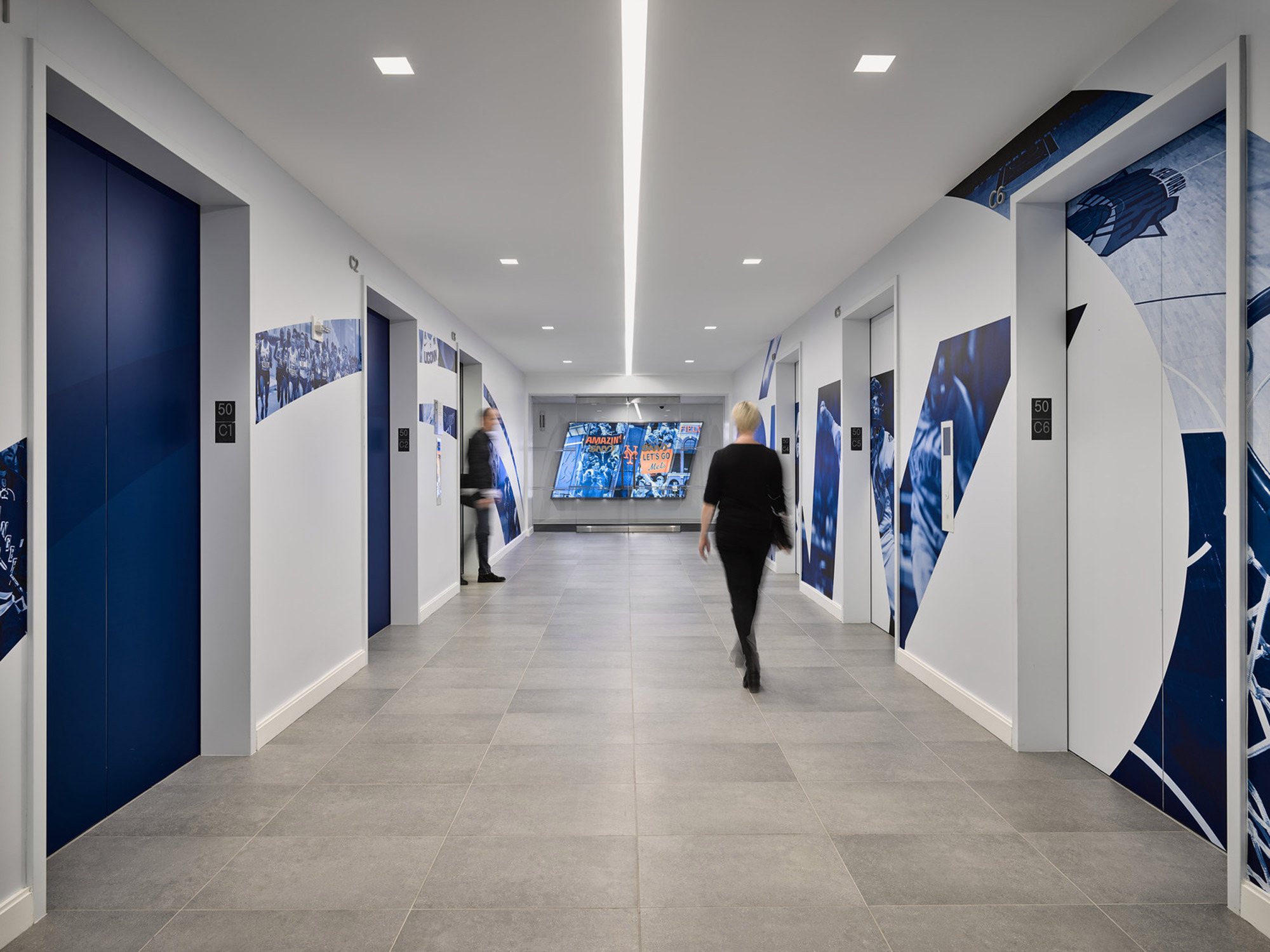 A modern corridor with polished grey floor tiles and white walls adorned by geometric patterns and vivid blue graphics. Mirrored doors interspersed with windows leading to offices, providing a blend of privacy and openness. A figure strides through, enhancing the sense of workplace dynamism.