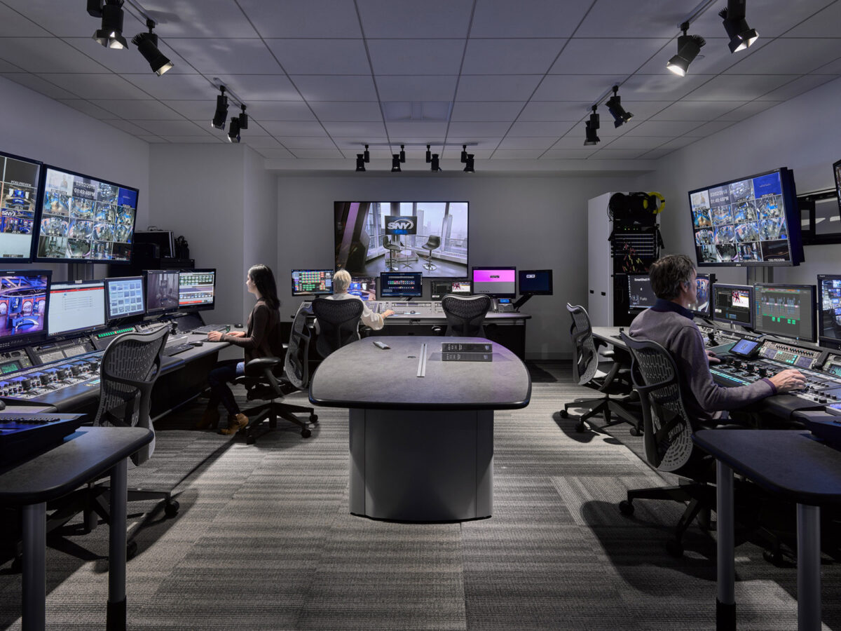 Modern control room with ergonomic workstations featuring multiple monitors set up on tiered desks for optimal viewing. Blue ambient lighting complements the sleek, dark-toned furniture, enhancing focus and reducing eye strain for technicians monitoring real-time data.