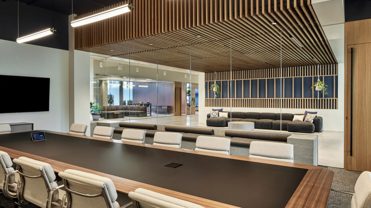 Modern conference room boasting sleek, dark-toned furnishings with a grand wooden table as the centerpiece, comfortable white chairs, pendant lighting above, and an inviting casual seating area against the backdrop of floor-to-ceiling windows revealing a cityscape.