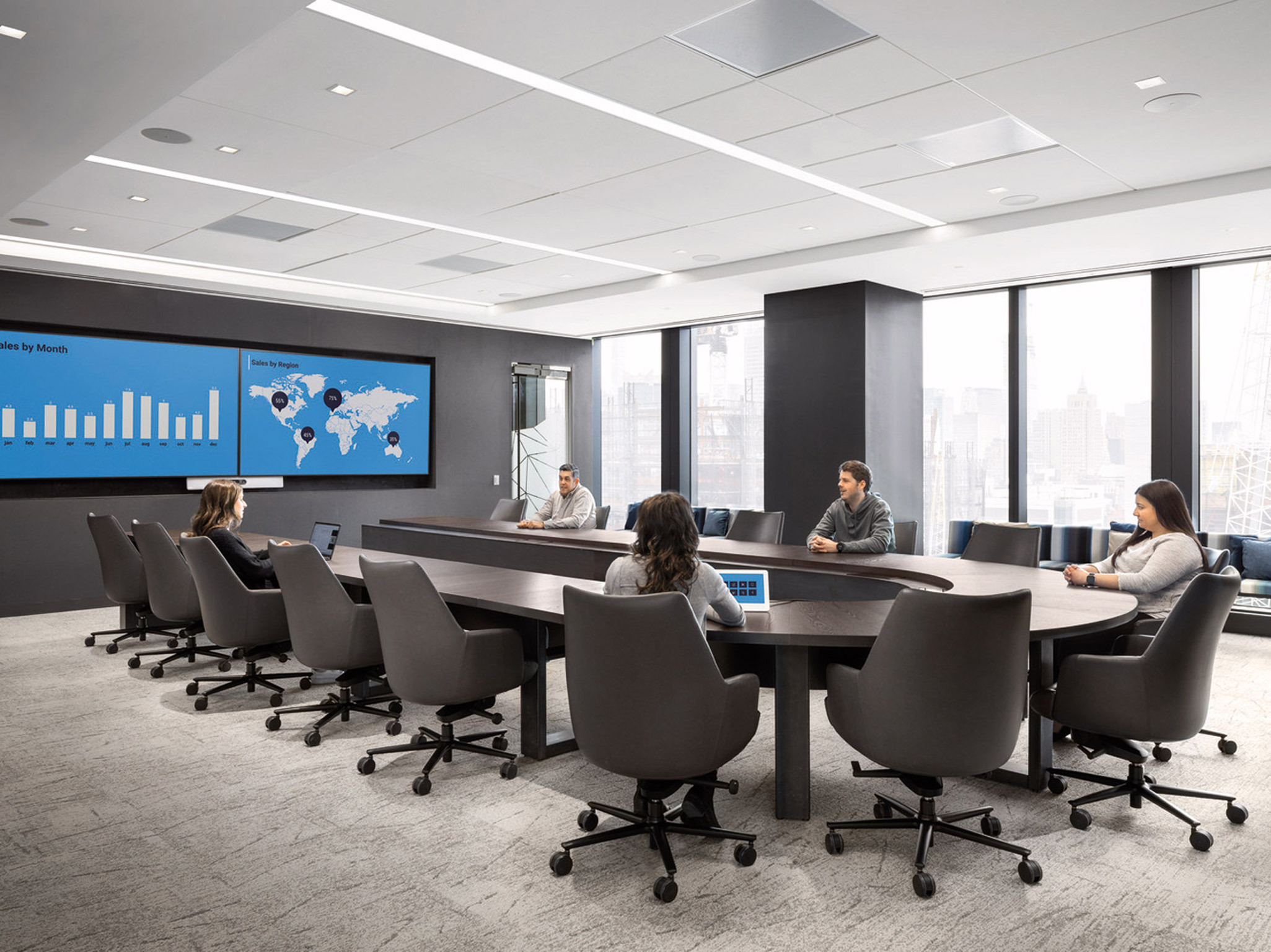 Modern conference room with a long oval table, ergonomic chairs, and a wall-mounted digital presentation screen. Natural light fills the room from floor-to-ceiling windows, complementing the subtle grey and blue color scheme.