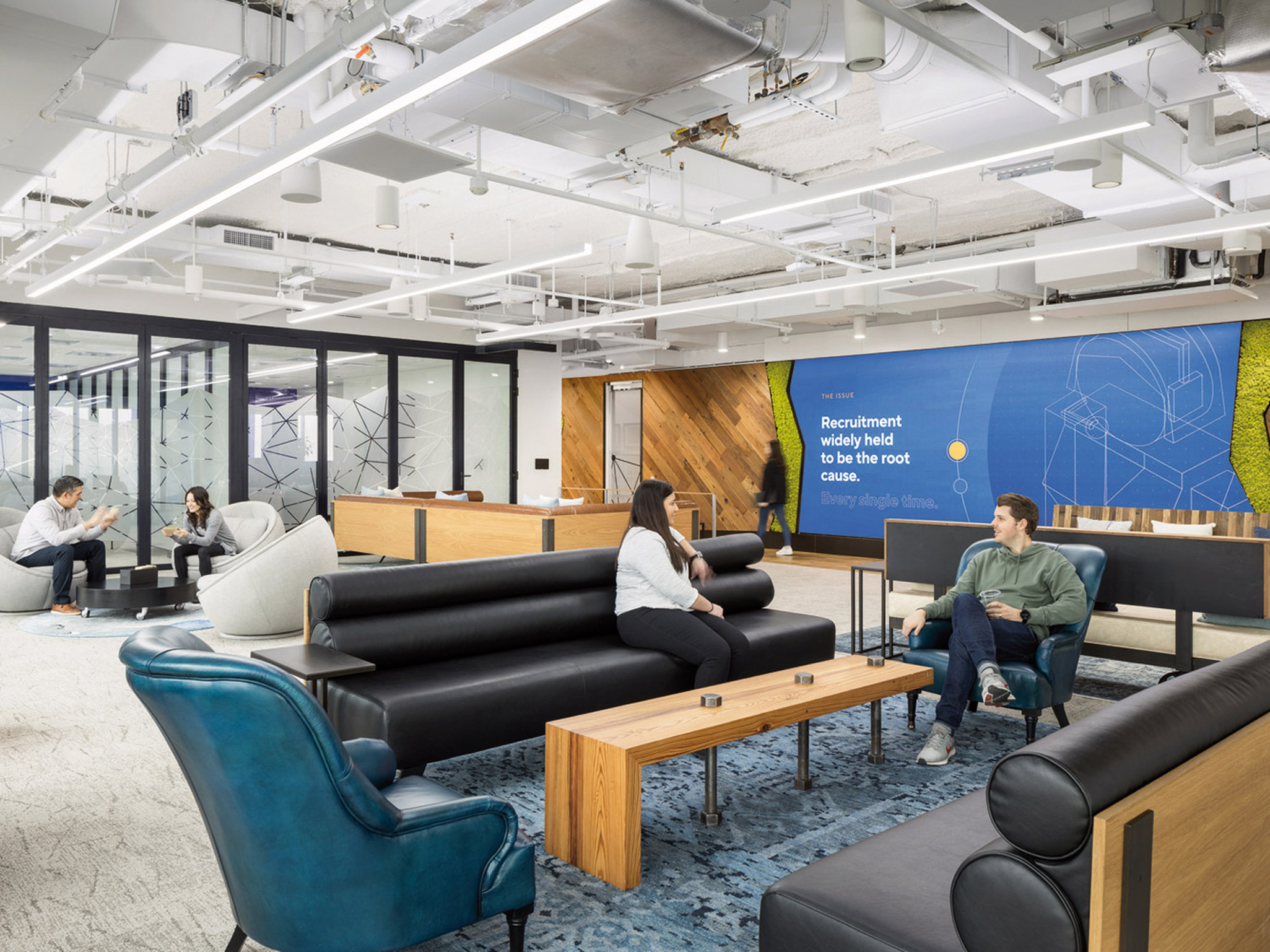 Modern office space with an open floor concept, featuring eclectic seating arrangements. Bold blue armchairs juxtapose with neutral-toned sofas, while a large recruitment-themed mural adds visual interest. Exposed ceiling beams and lighting fixtures create an urban industrial ambiance.