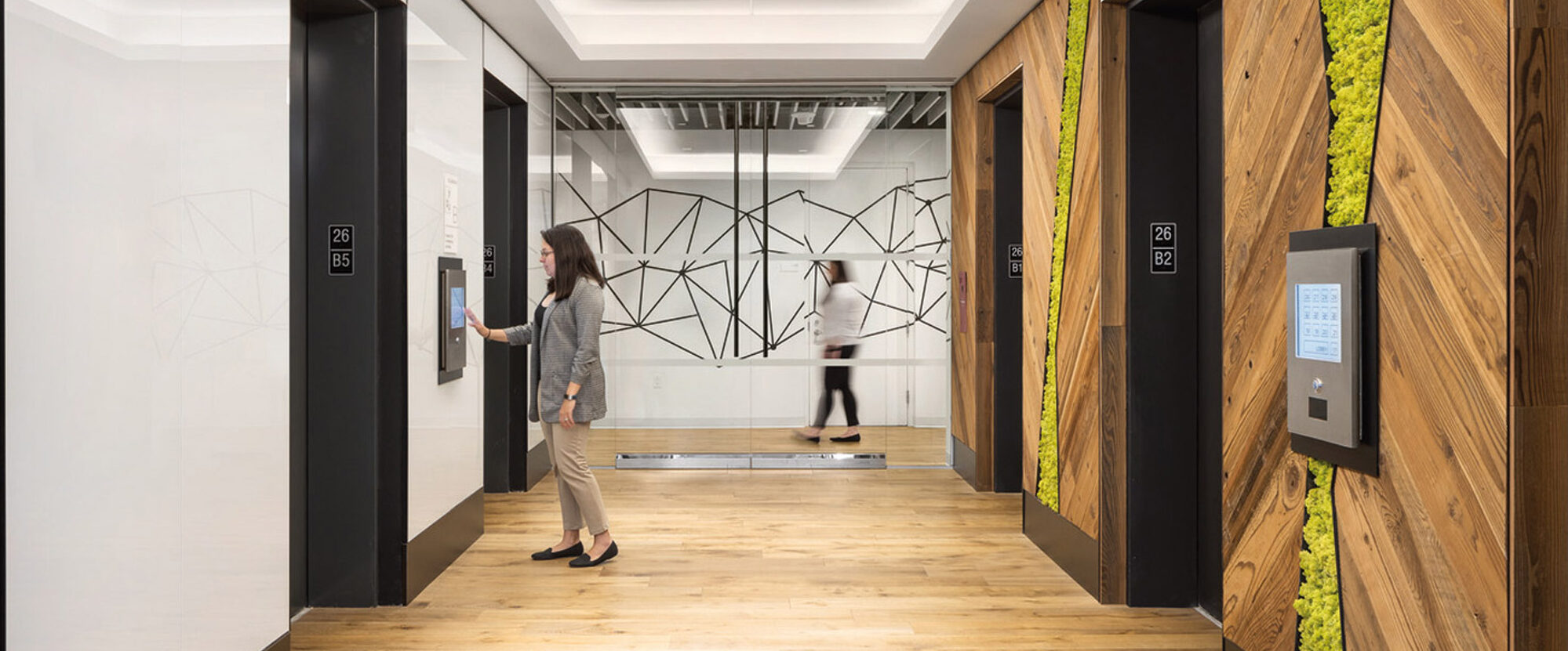 Modern office hallway featuring warm wood floors, contrasting white and woodgrain walls with vibrant green moss accents, geometric patterns, and sleek elevator doors; inclusive design with accessible signage and an interactive directory panel.