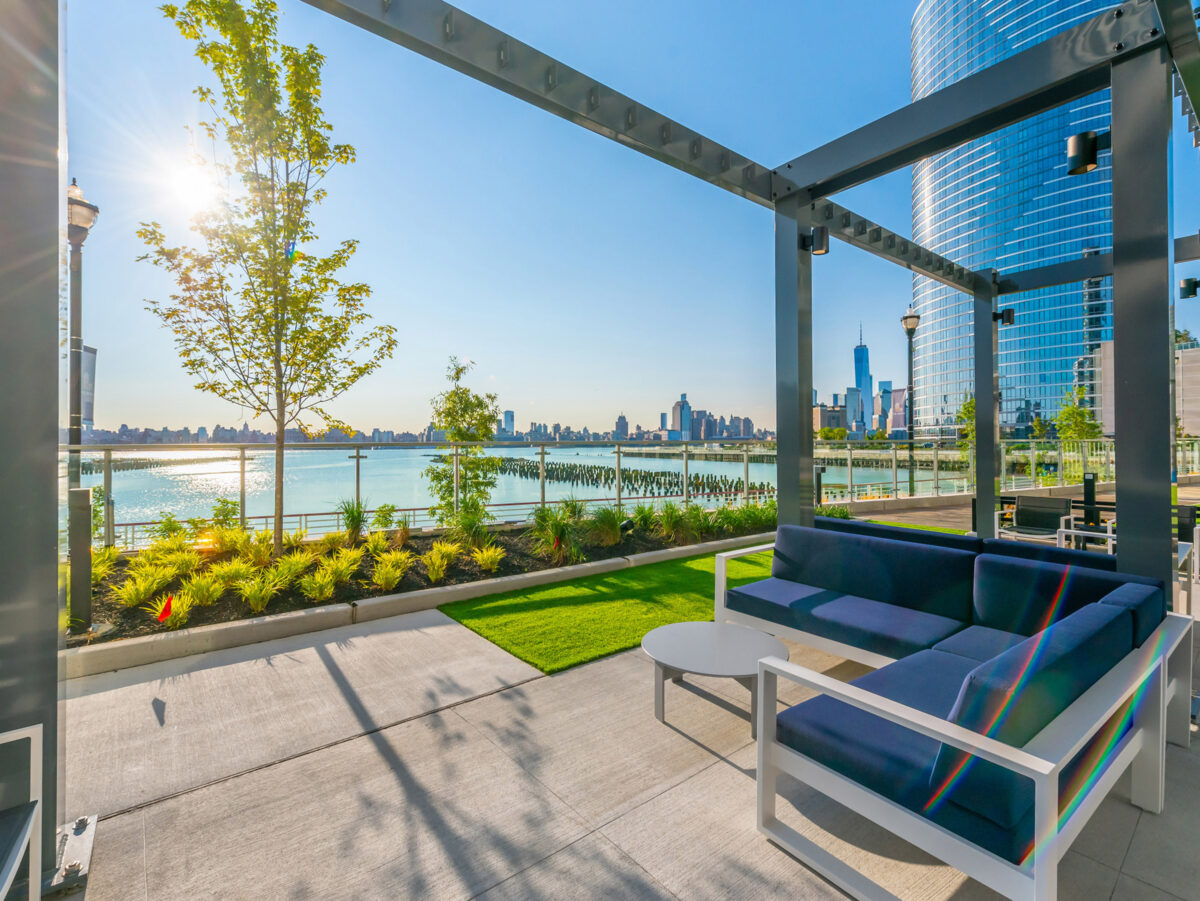 Modern outdoor lounge area featuring sleek white sofas with colorful cushions, set on green turf, encompassed by a high-rise cityscape background and water views, under a metal pergola framing the bright blue sky.