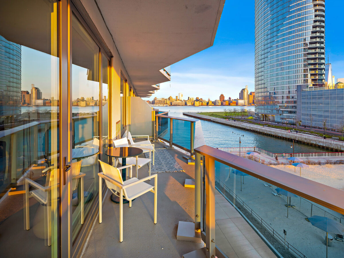 Modern balcony with sleek white furniture overlooking an urban skyline during sunset. The glass balustrade provides an unobstructed view, enhancing the seamless indoor-outdoor transition.