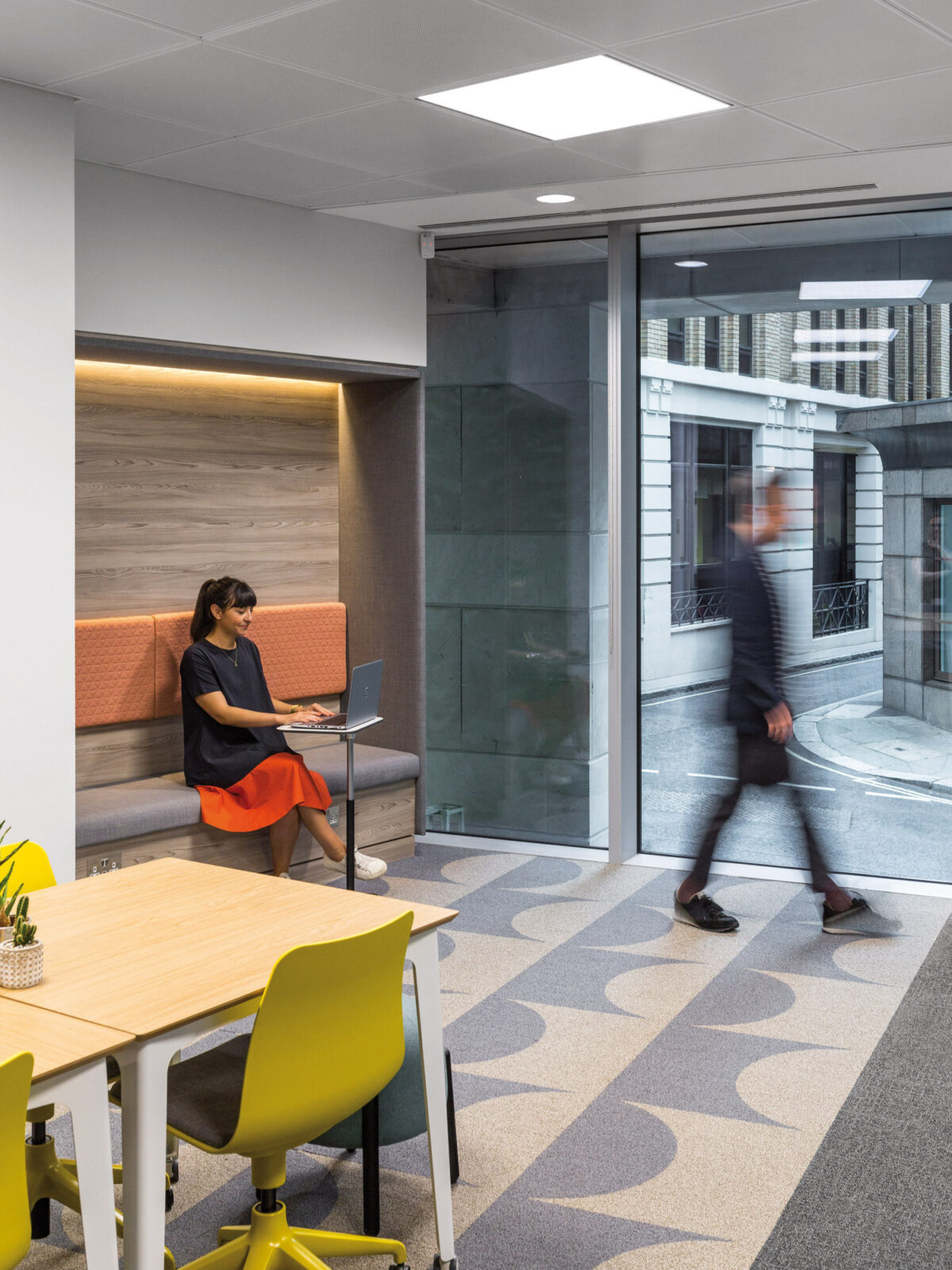 Modern office breakout space featuring a woman seated at a wall-mounted desk, warm wood paneling, vibrant orange upholstered seating, and geometric-patterned gray carpet. A blurred figure walking past the glass partition highlights the active work environment.
