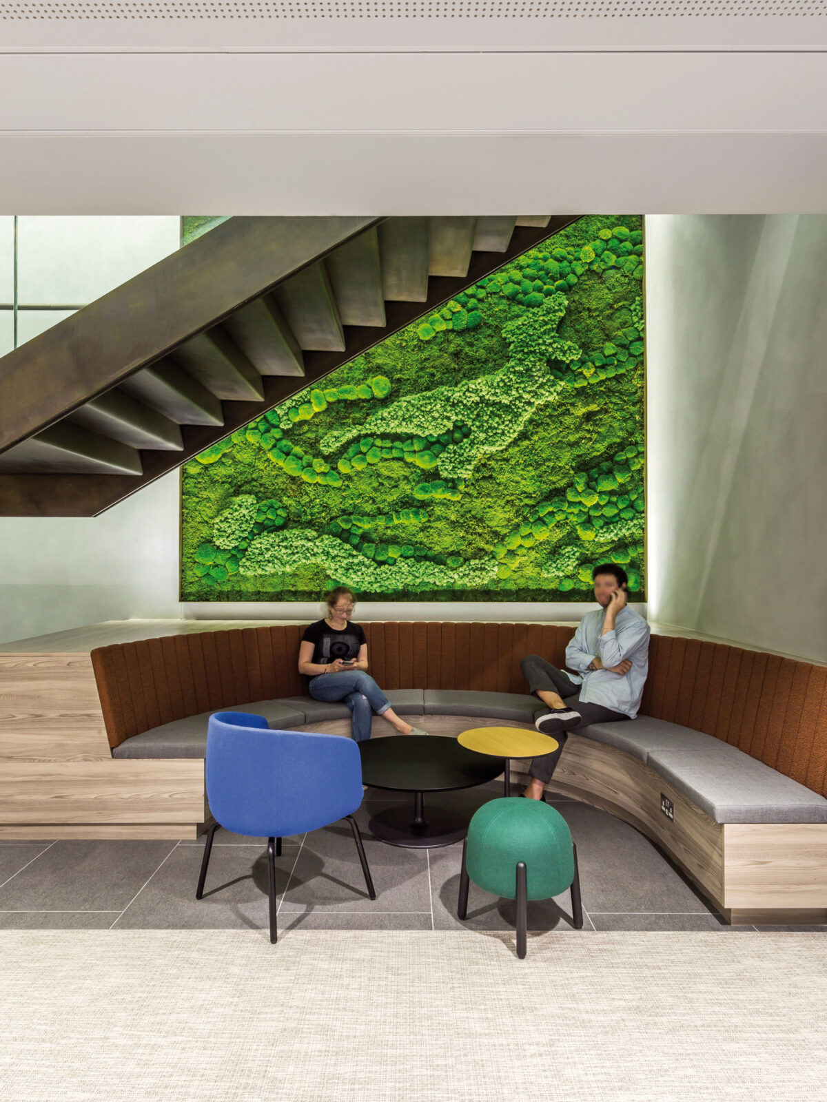 Modern stairwell adjacent to an expansive green moss wall. Below, a built-in wooden bench curves around the room, complemented by vibrant blue and yellow accent chairs and a round table, creating a dynamic waiting area.