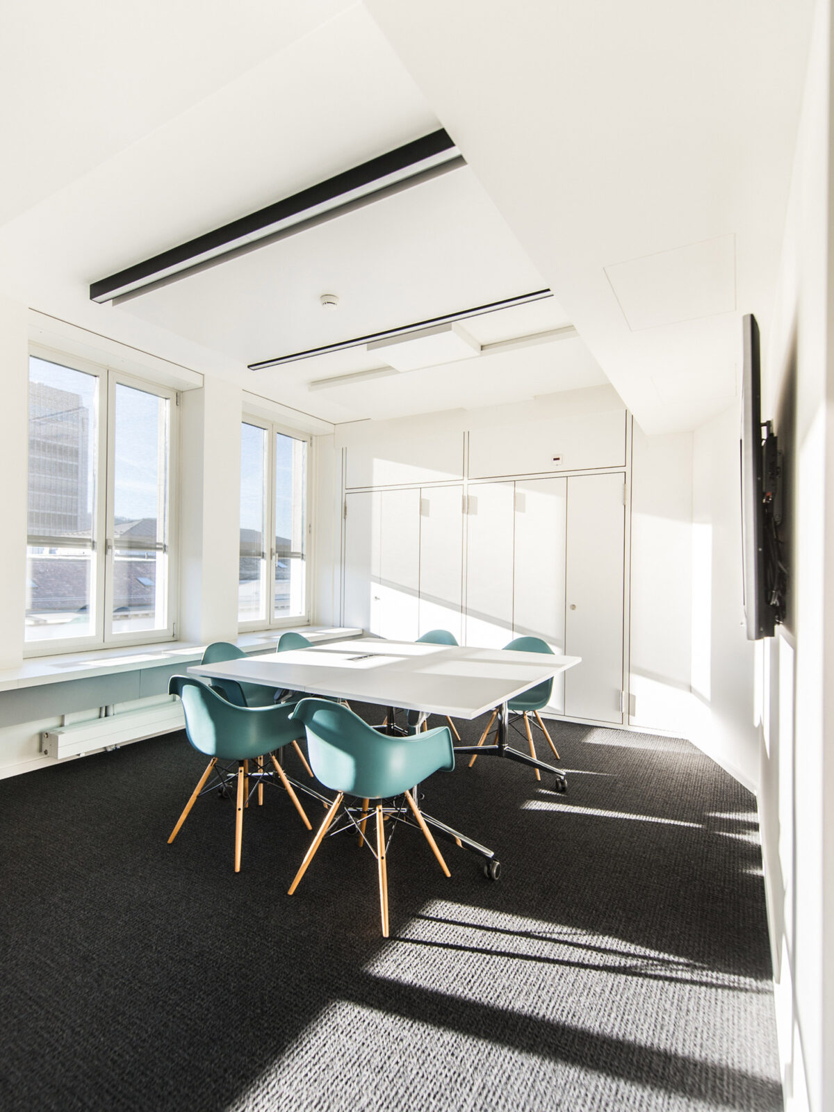 Minimalist conference room bathed in natural light, featuring a sleek white table, turquoise Eames-style chairs, dark textured carpeting, and a discreet ceiling-mounted projector.