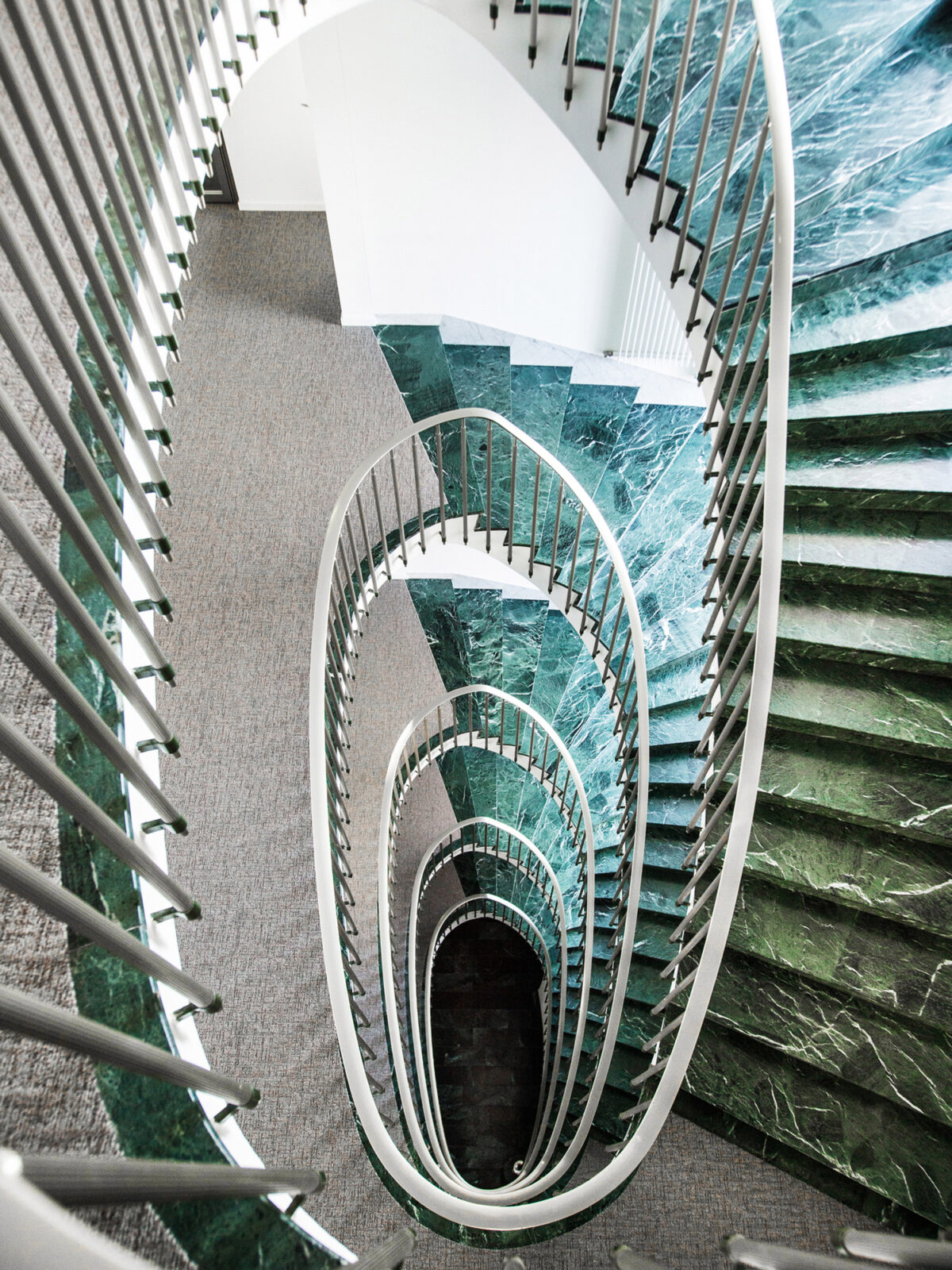 Spiral staircase with white balustrades and steps; below, a patterned emerald floor gives an impression of depth, enhancing the sculptural quality of the architecture.