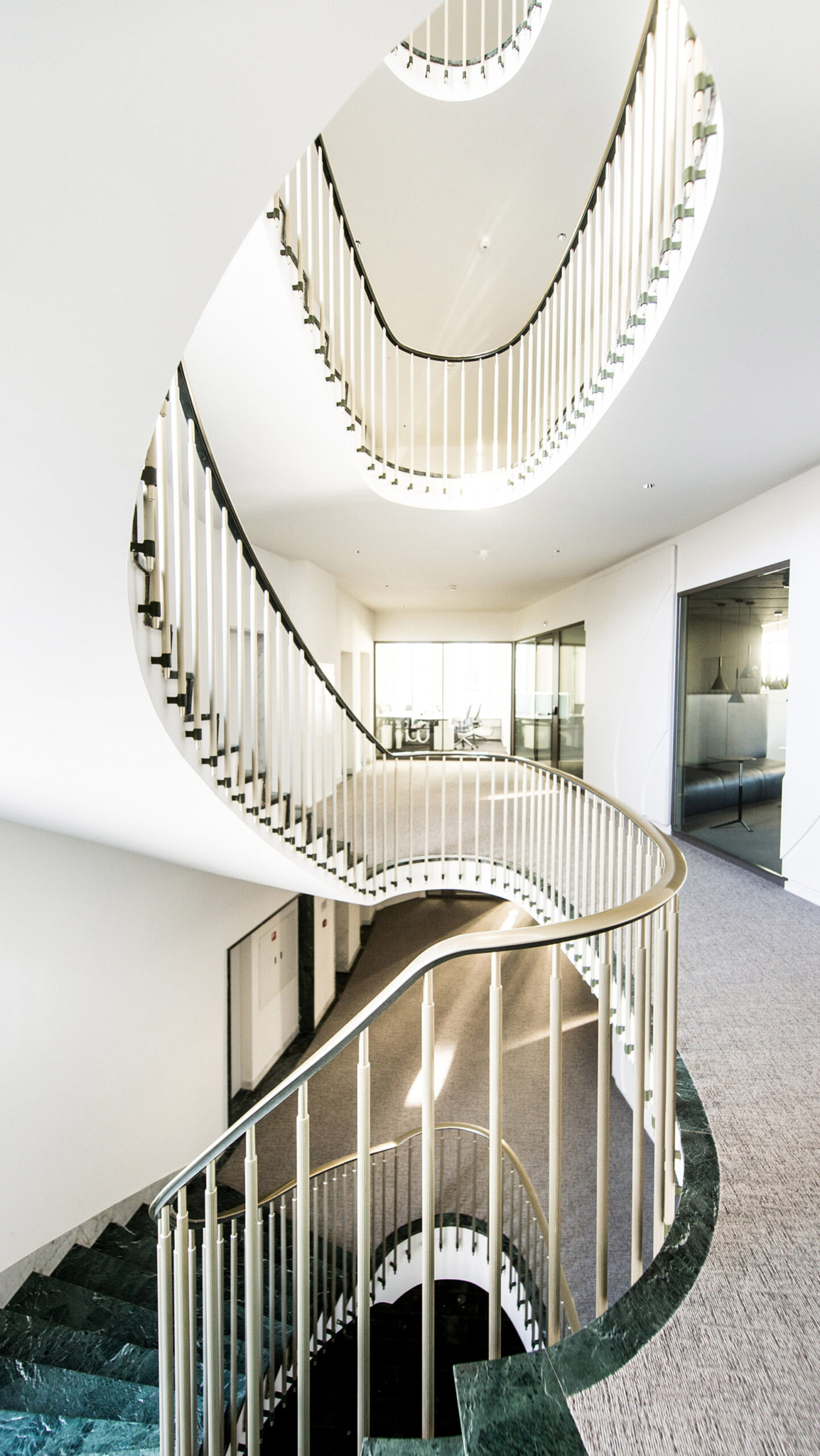 Elegant spiral staircase with white balustrades creating a visually striking contrast against the dark polished stone flooring. The design offers a seamless flow through the levels, complemented by abundant natural light reflecting off the crisp, clean lines.