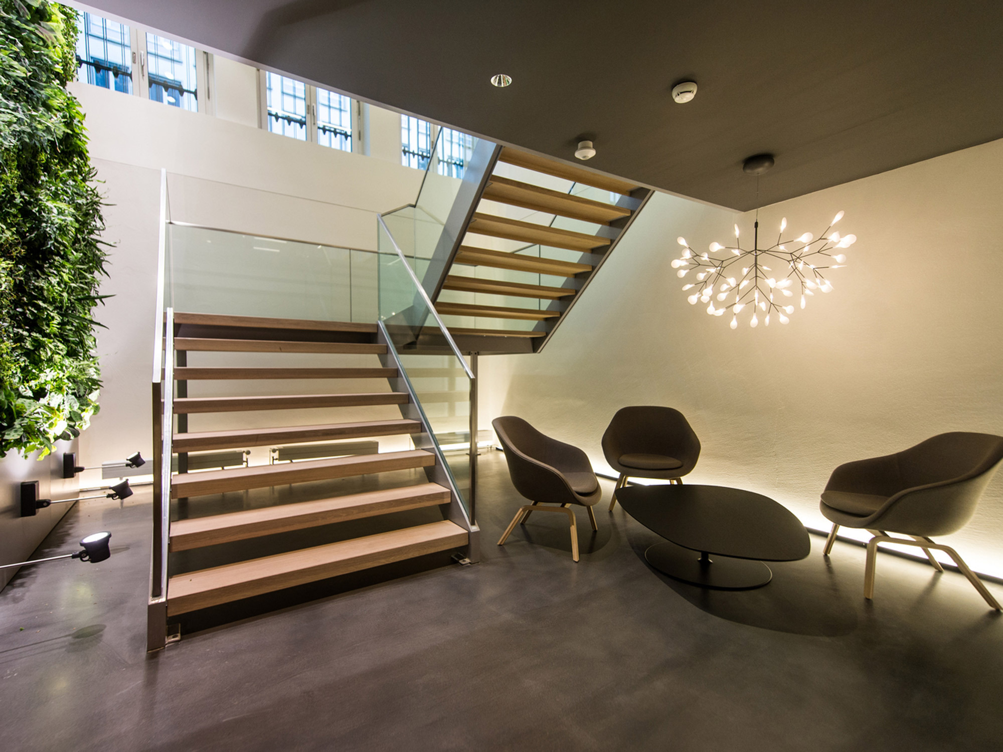 Modern staircase in a contemporary space with floating wooden steps, glass balustrades, and a vertical garden, complemented by minimalist seating and a statement lighting fixture.