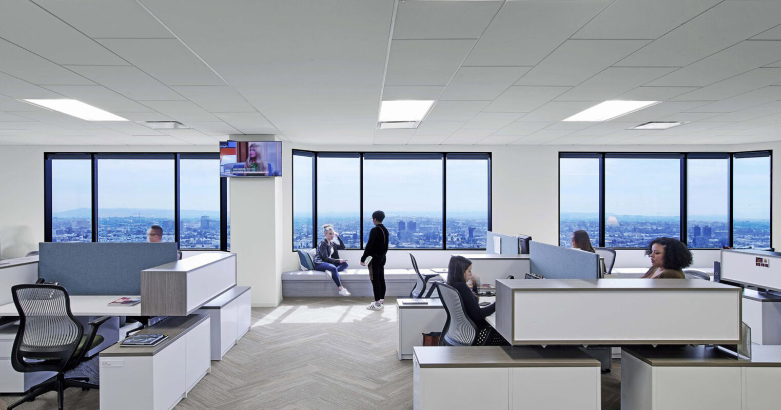 Modern office space with ergonomic workstations, carpeted floors, and expansive windows offering an urban skyline view. Collaborative seating areas are interspersed, enhancing the open-plan layout. Ambient lighting complements the natural daylight, fostering a productive work environment.