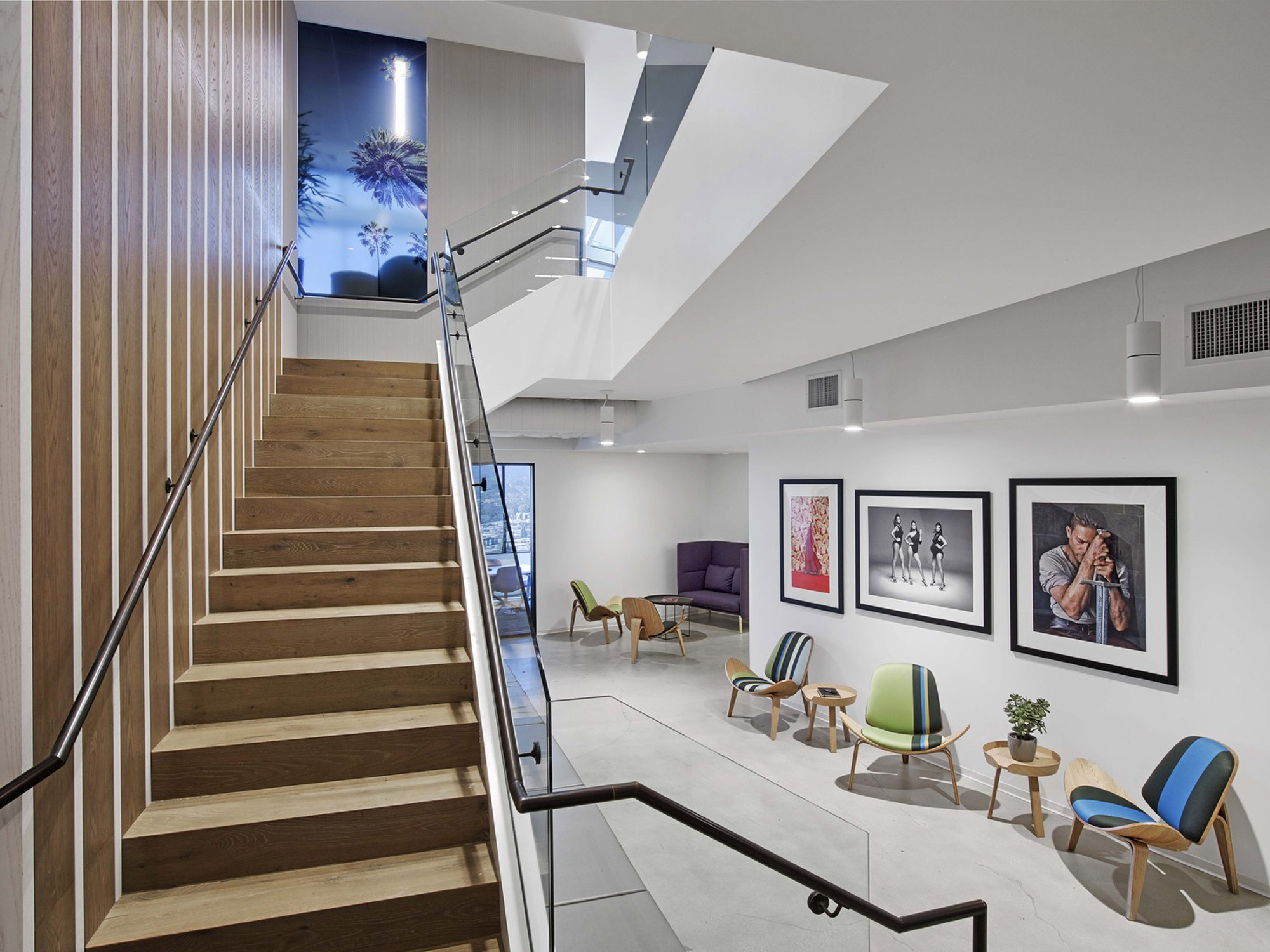 This interior features a modern staircase with warm wooden steps flanked by a sleek metal handrail, leading up to a brightly lit upper level. Below, a seating area boasts a mix of colorful mid-century modern chairs, with gallery-style framed artwork adorning the white walls.