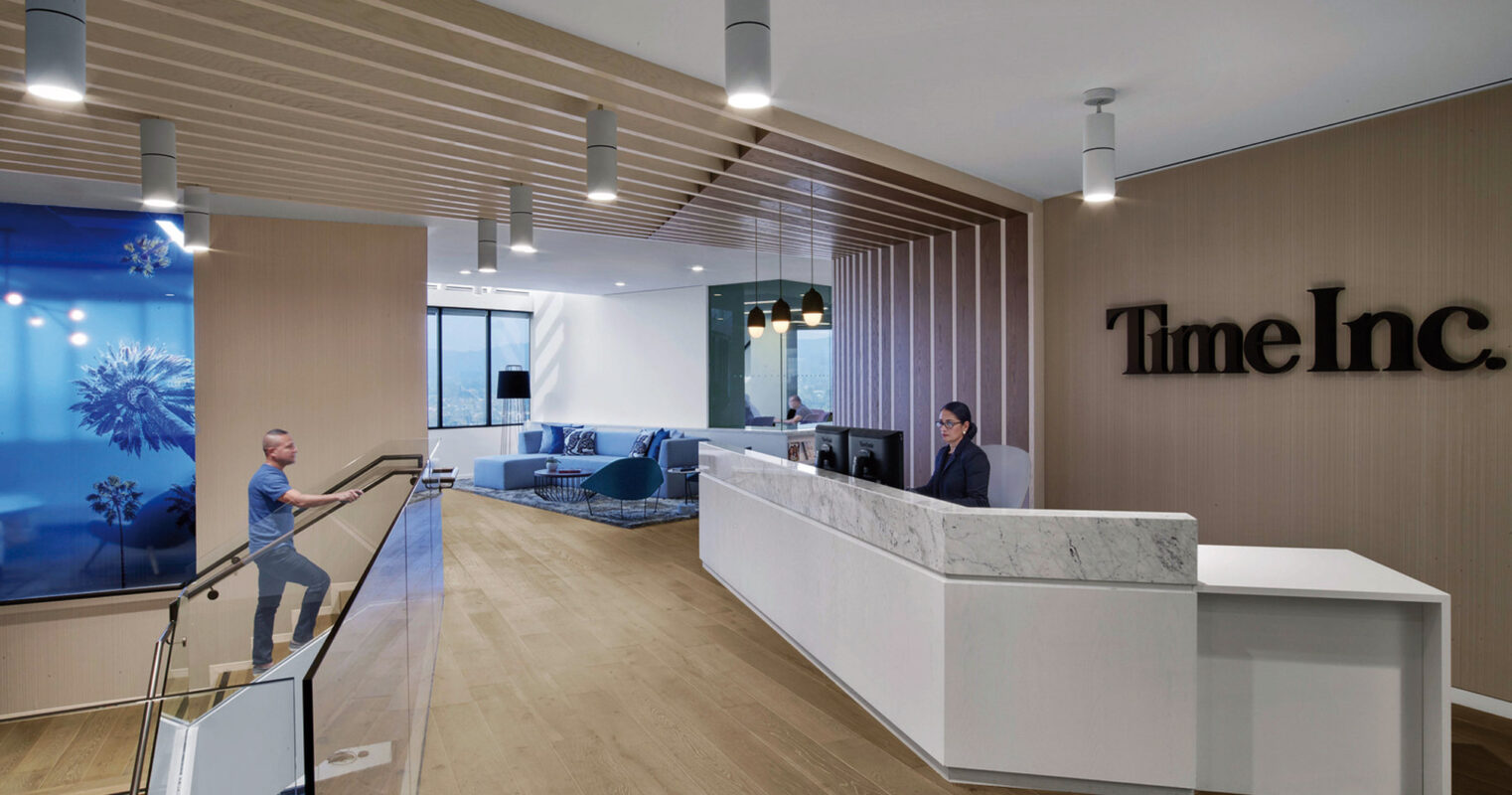 Modern office lobby featuring a sleek white reception counter with the Time Inc. logo on a wooden slat wall. Blue-toned artwork adds a pop of color, complemented by pendant lighting and contemporary seating area in the background.