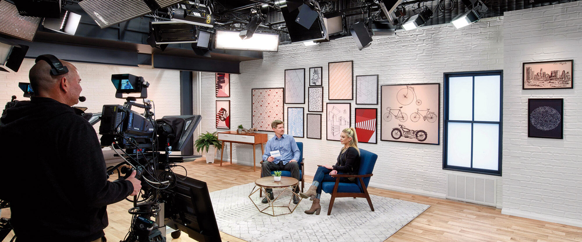 Contemporary television studio with two guests seated on minimalist furniture, under focused lighting, with a textured white brick wall adorned with assorted framed artwork in the background.