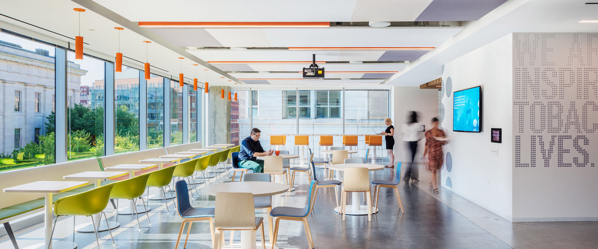 Bright, modern office break area with floor-to-ceiling windows, offering ample natural light. White tables with colorful chairs in green and blue hues complement the vibrant orange light fixtures overhead. Interactive LED screen enlivens the space, surrounded by dynamic text wall art, fostering an engaging work environment.