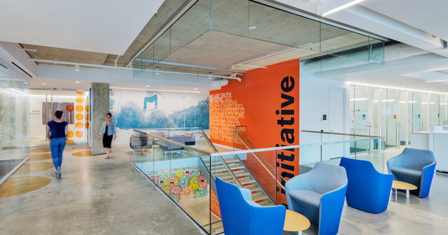 Modern office lobby with vibrant orange accent wall featuring typographic art, complemented by sleek glass stair railing. Blue upholstered armchairs add a pop of color, set against the crisp white walls and polished concrete floor. Natural light enhances the space's open, airy ambiance.