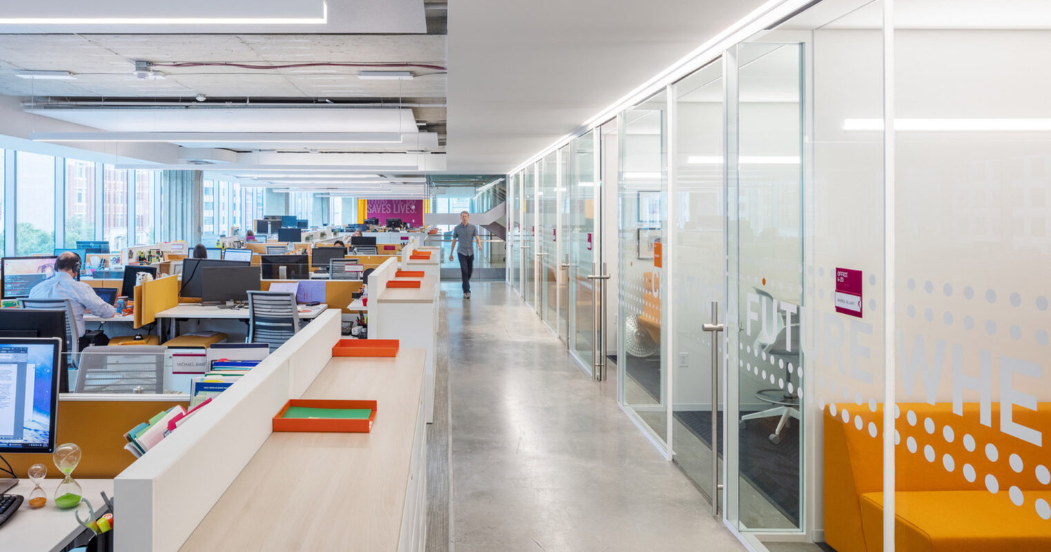 Open-plan office space showing a clear distinction between collaborative areas with clusters of desks and private glass-walled meeting rooms, featuring bright pops of color and ergonomic furniture. Natural light fills the space, complementing the contemporary and functional design.