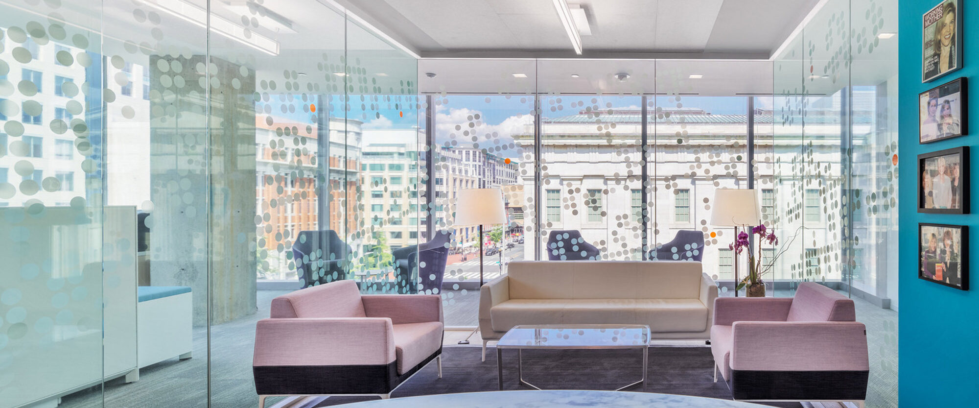 Modern office reception area with floor-to-ceiling windows providing natural light, two blush armchairs, a beige sofa atop a marble rug, and a frosted glass room divider with circular patterns. Art pieces adorn the adjacent wall, harmonizing with the city view beyond.