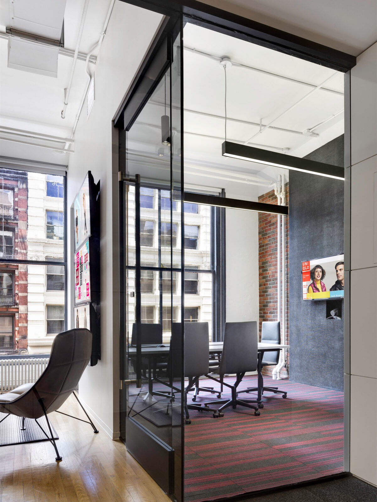 Modern office space highlighted by natural light, featuring a glass partition, sleek black frame, contemporary furnishings, and an exposed brick wall, complemented by a vibrant red patterned area rug.