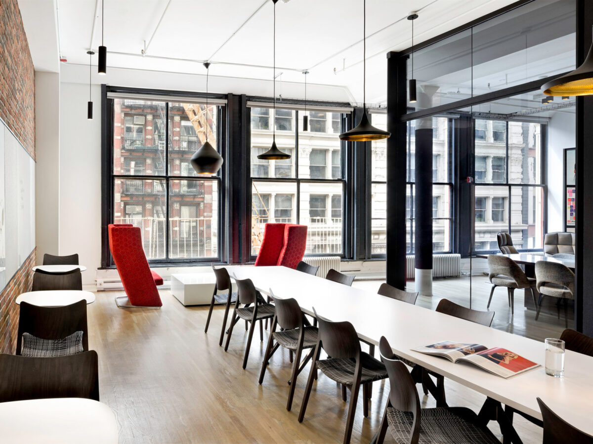 Spacious industrial-style loft features high ceilings and large windows offering ample natural light. Exposed brick wall adds texture, complemented by sleek pendant lighting. A long, white communal table with a mix of black chairs and red high-back seats anchors the room, promoting collaboration and interaction.