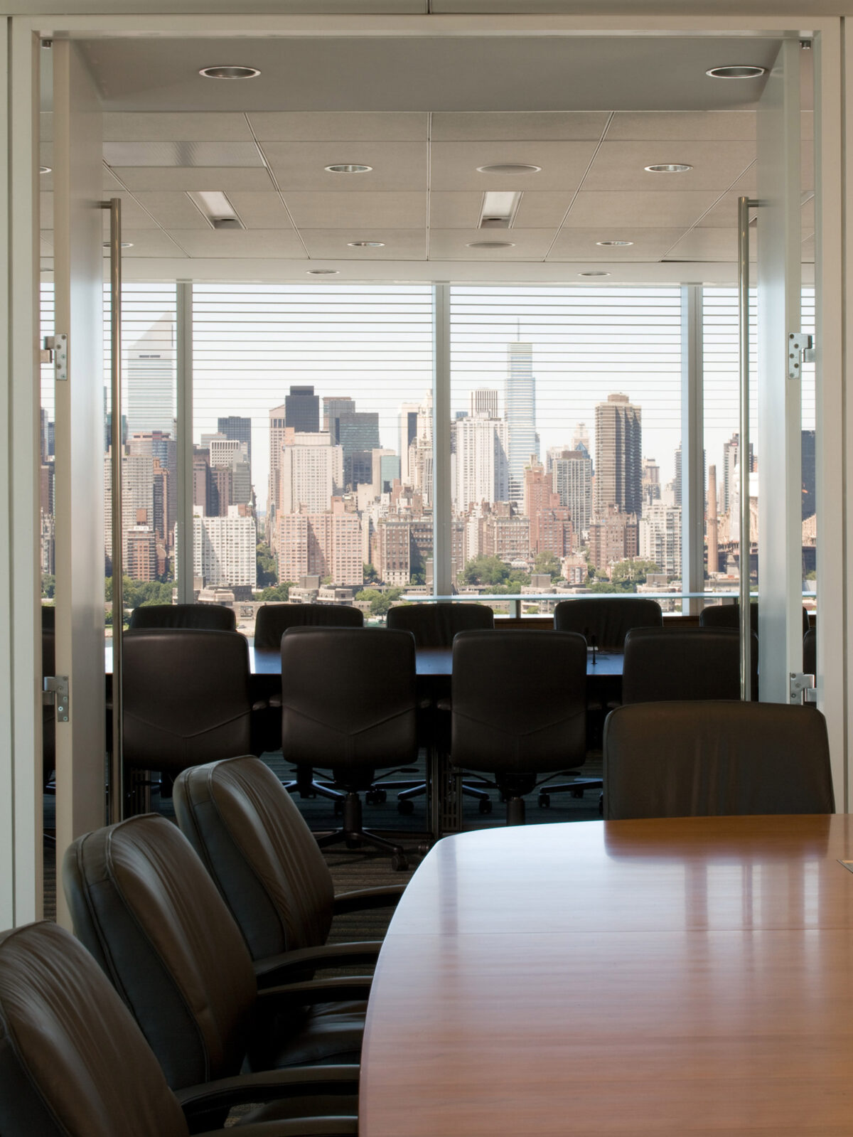 Modern boardroom with sleek, black rolling chairs around a long, polished wooden table, featuring expansive windows that frame a panoramic city skyline view, accentuating the room's natural lighting and urban context.