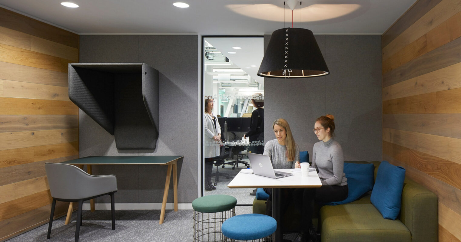 Modern office lounge with neutral gray walls accented by natural wooden paneling. Includes a plush corner sofa, sleek black pendant light, and ergonomic chairs. The space fosters collaboration with a central meeting table and combines comfort with contemporary style.