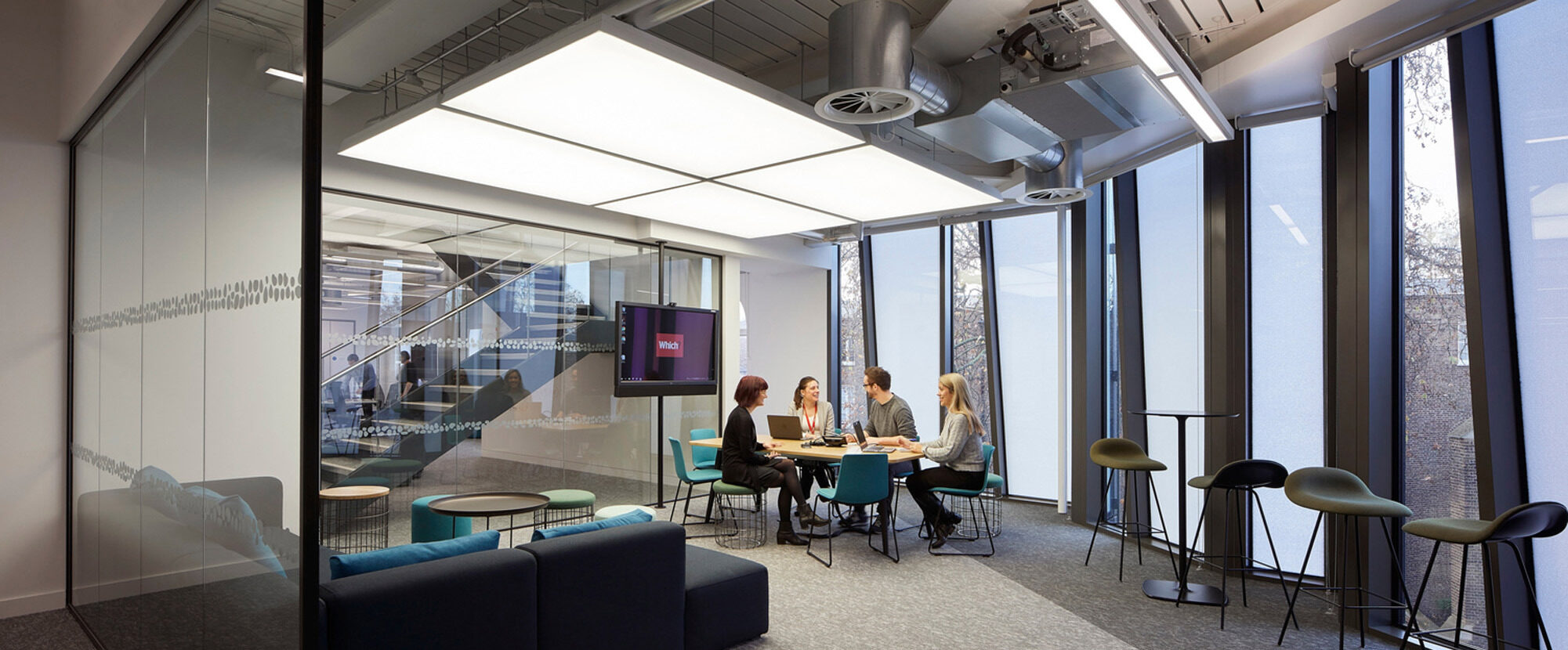Modern office meeting area with expansive windows, natural light, and a sleek glass partition. Sleek bar stools complement a high table, while a cozy blue sofa encircles a lower wooden table, fostering a collaborative atmosphere.