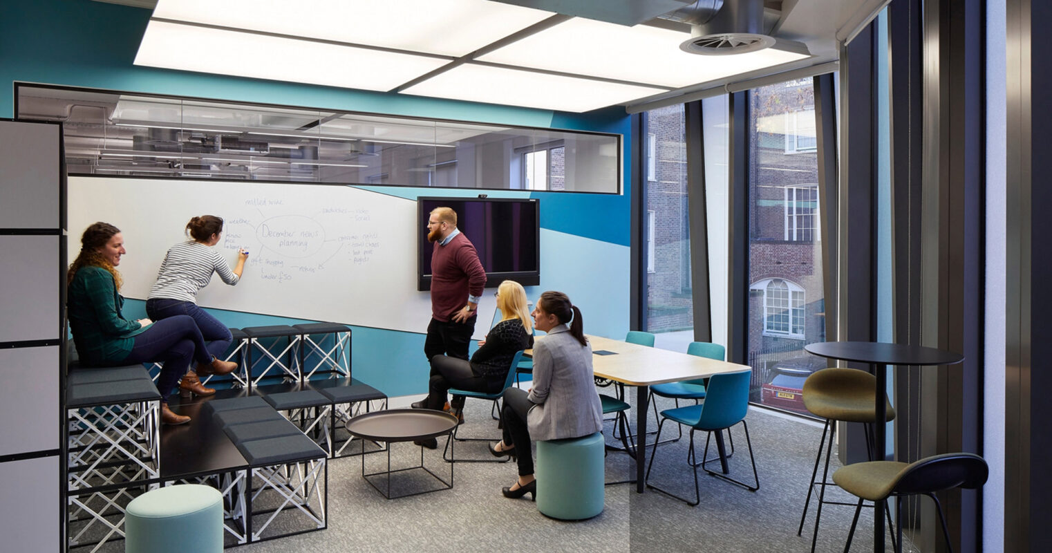 Modern office space featuring vibrant blue partitions, whiteboard for collaborative brainstorming, diverse seating options from poufs to ergonomic stools, and ample natural light complementing artificial task lighting. The design fosters a dynamic and interactive work environment.