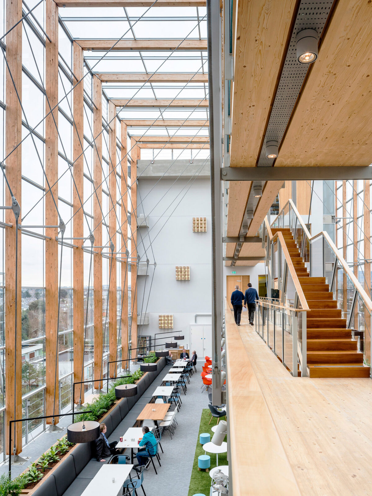 Spacious atrium with soaring wooden columns flanking a central walkway. Natural light floods the two-story space, highlighting the warm wood paneling and staircases that lead to balconies overlooking modern, open-plan seating areas below.