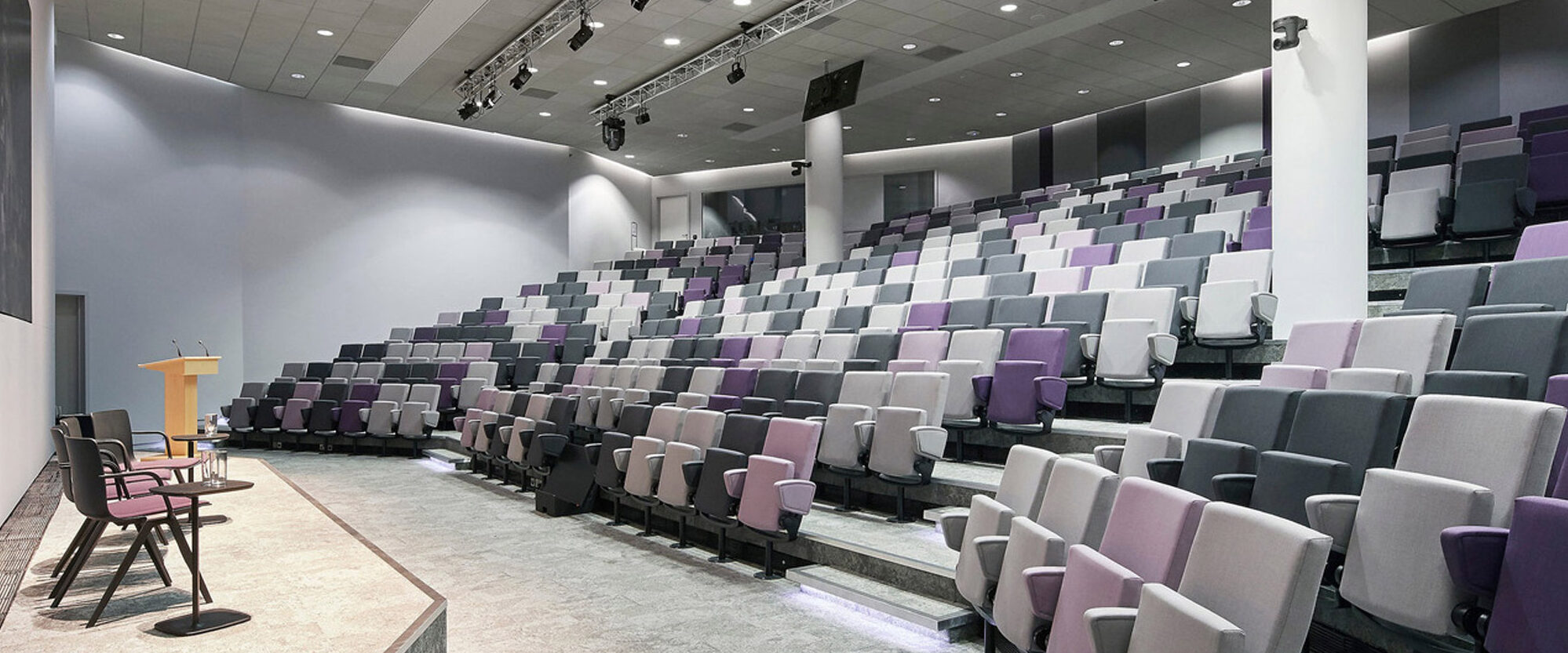 Contemporary auditorium with tiered seating featuring shades of gray and purple upholstered chairs, focused lighting from ceiling spots and ambient wall fixtures, and a central stage with a wooden podium flanked by chairs.