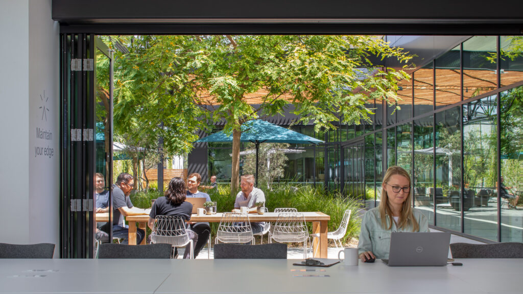 Professionals gather at a modern outdoor meeting space featuring minimalistic furniture and lush greenery, with a transparent glass facade integrating the interior office environment and the vibrant outdoor setting.