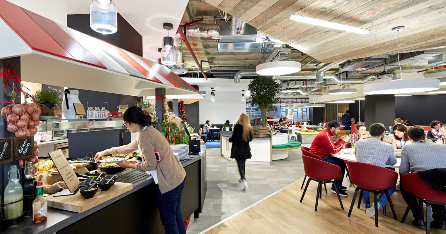 Modern office cafeteria with industrial ceiling elements, exposed ductwork, and pendant lighting. Wooden beams accentuate the ceiling, contrasting with sleek red and black seating. Employees enjoy meals at a central buffet island.