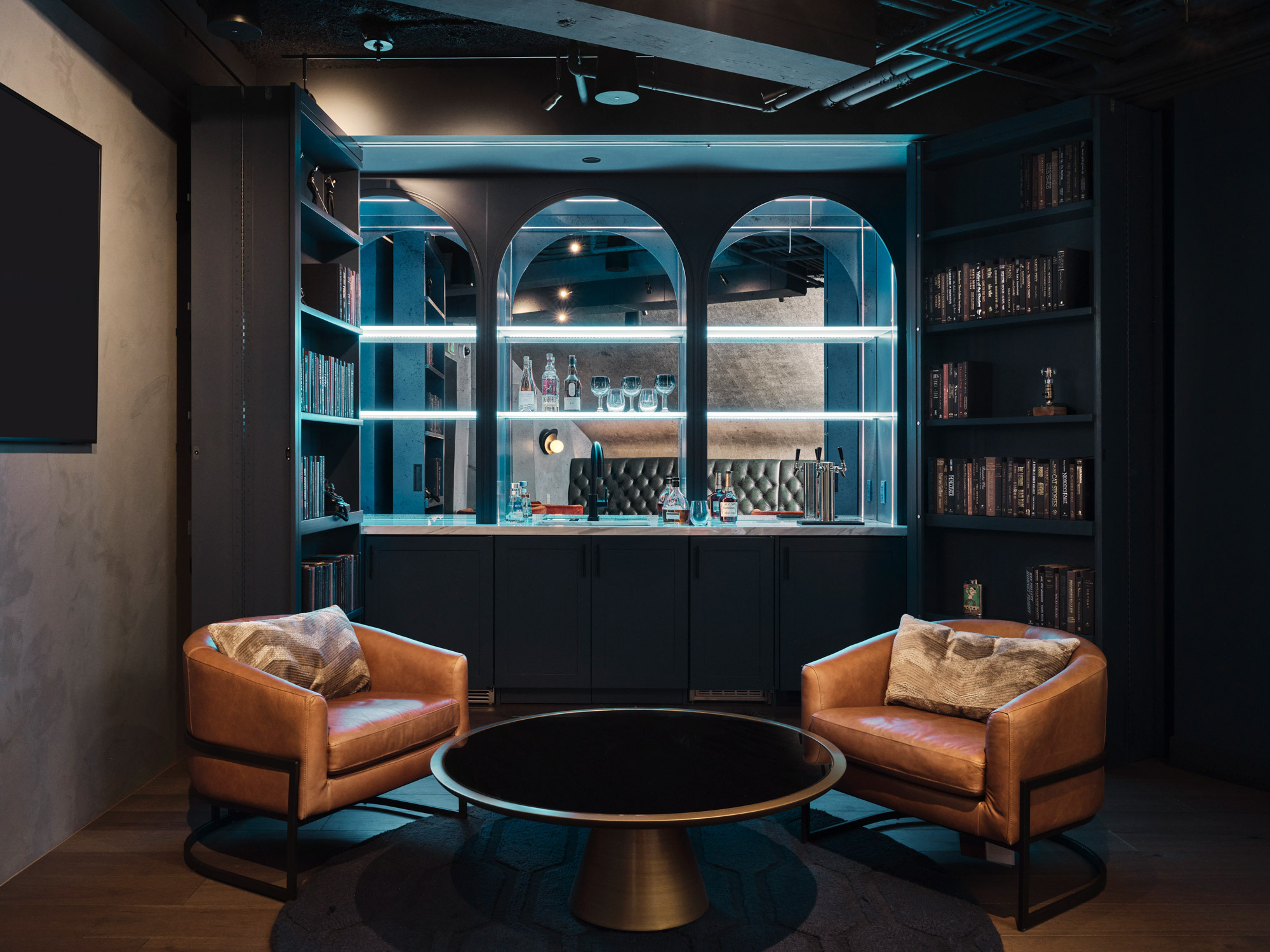 Modern lounge with two caramel leather armchairs flanking a circular black coffee table, set against a backdrop of deep blue bookshelves with integrated lighting, showcasing a curated collection of books and decorative objects. The shelves feature elegant arch-top compartments, enhancing the sophisticated, intimate ambiance.