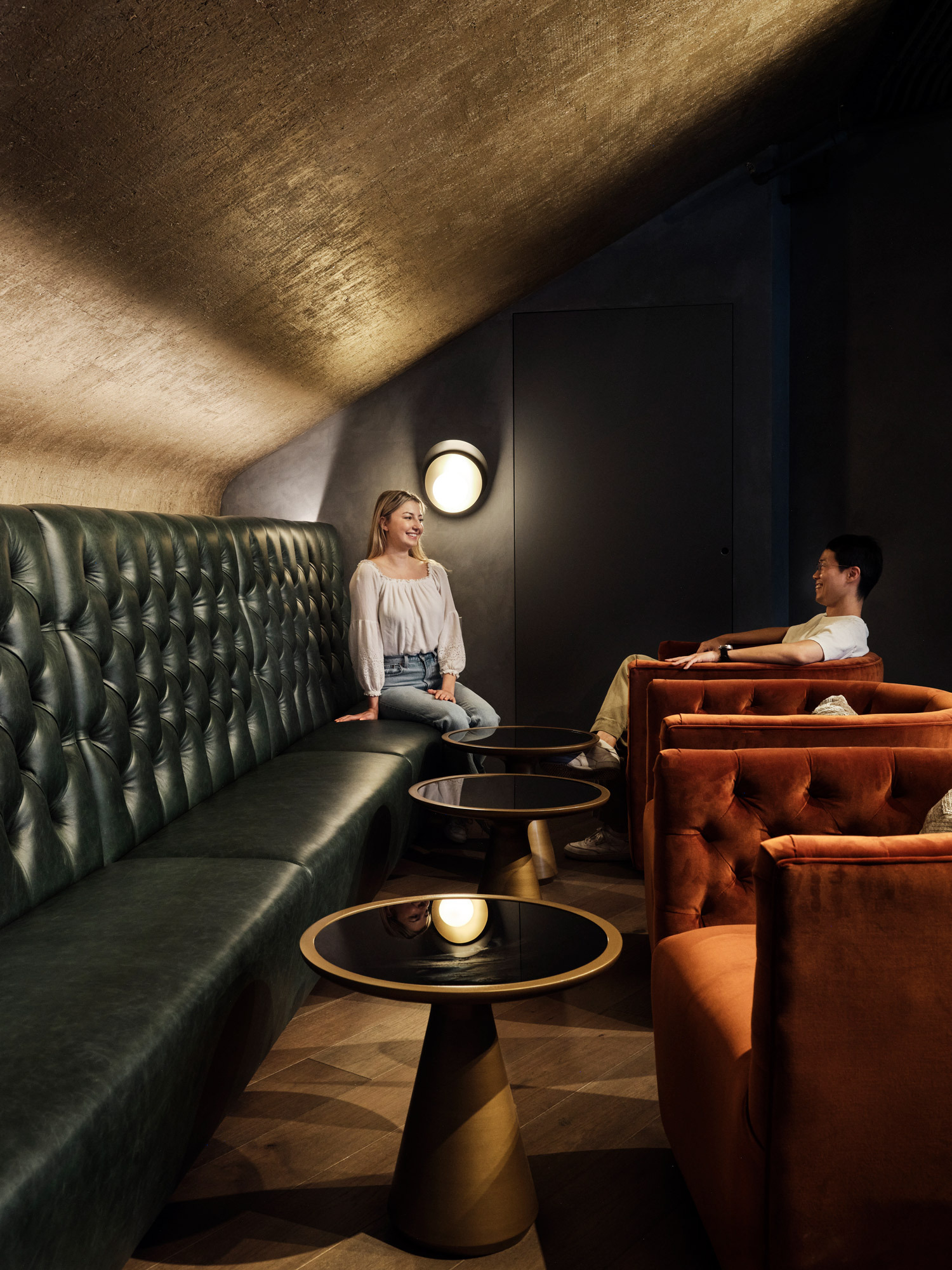 Soft lighting accentuates the luxurious textures within this upscale lounge, where richly tufted emerald green leather booths contrast with sumptuous terracotta armchairs, and sleek gold-toned side tables provide a sophisticated touch amidst the moody ambiance.