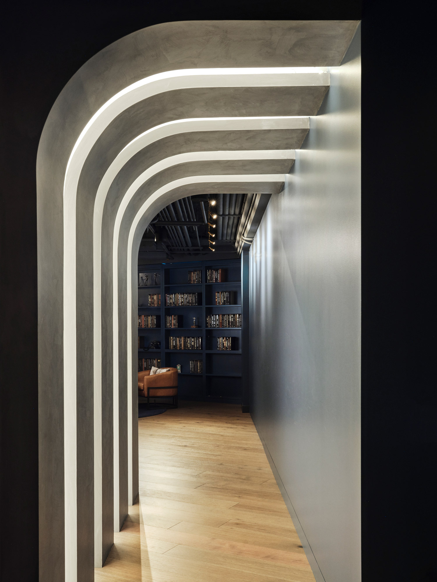 A sequence of arches with layered lighting creates a dynamic hallway contrasted against a dark-toned bookcase-lined room, leading the eye through the space with organic movement, highlighting fluidity and depth in contemporary interior design.
