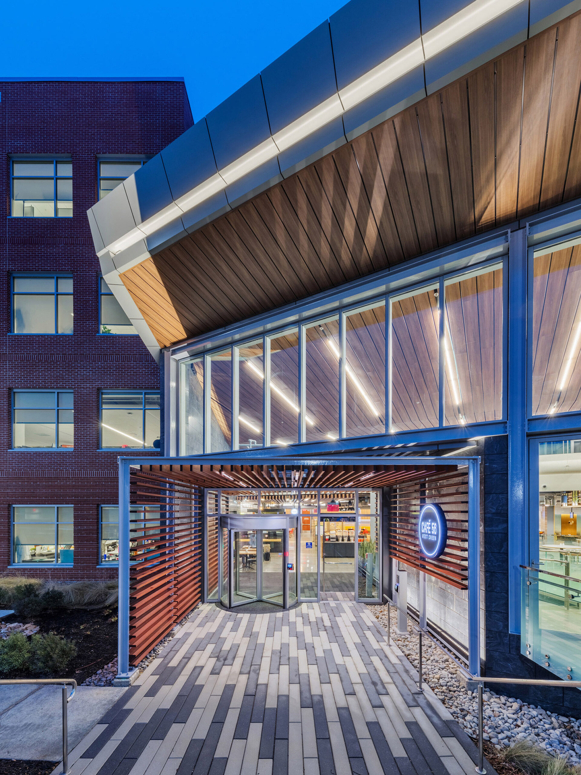 Modern architectural design featuring a building with distinctive brickwork and a curved, wood-paneled overhang leading to a glass entrance, illuminated by sleek, exterior lighting at twilight.