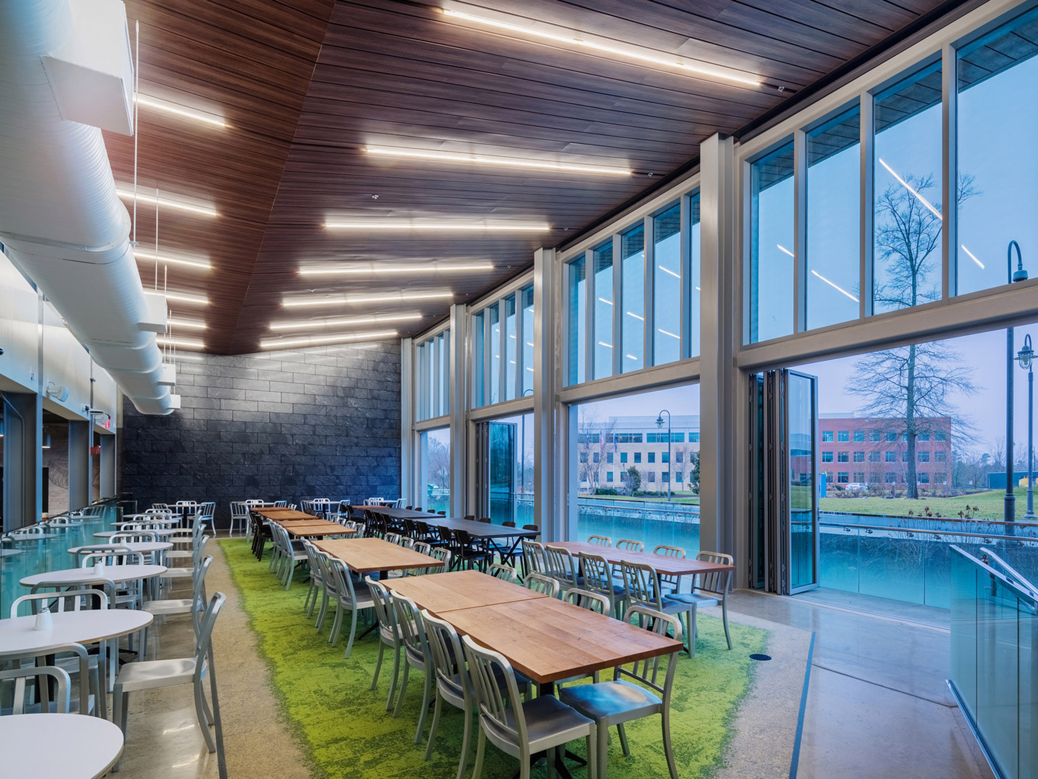 Spacious dining hall boasting high ceilings with wooden slats, wall-to-wall glazing providing ample natural light, and modern rectangular tables on a vibrant green carpet, complementing the exterior view of serene waters and architecture.