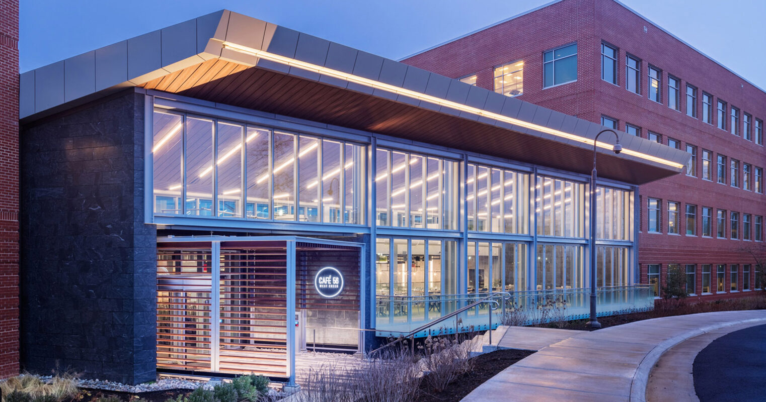 Modern educational building facade at twilight featuring a glass-walled walkway, illuminated interior spaces, sleek metal roof accents, and warm wooden cladding on a revolving entrance door area. The design integrates clear and opaque materials harmoniously, showcasing contemporary architectural style.