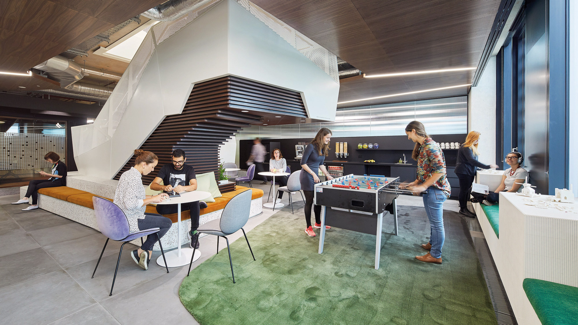 Modern open-plan office space with a variety of seating arrangements featuring eclectic furniture. Employees engage casually around a foosball table. Overhead, exposed ductwork complements the industrial-chic aesthetic, while vibrant green carpeting adds a pop of color, aiding in zone delineation.