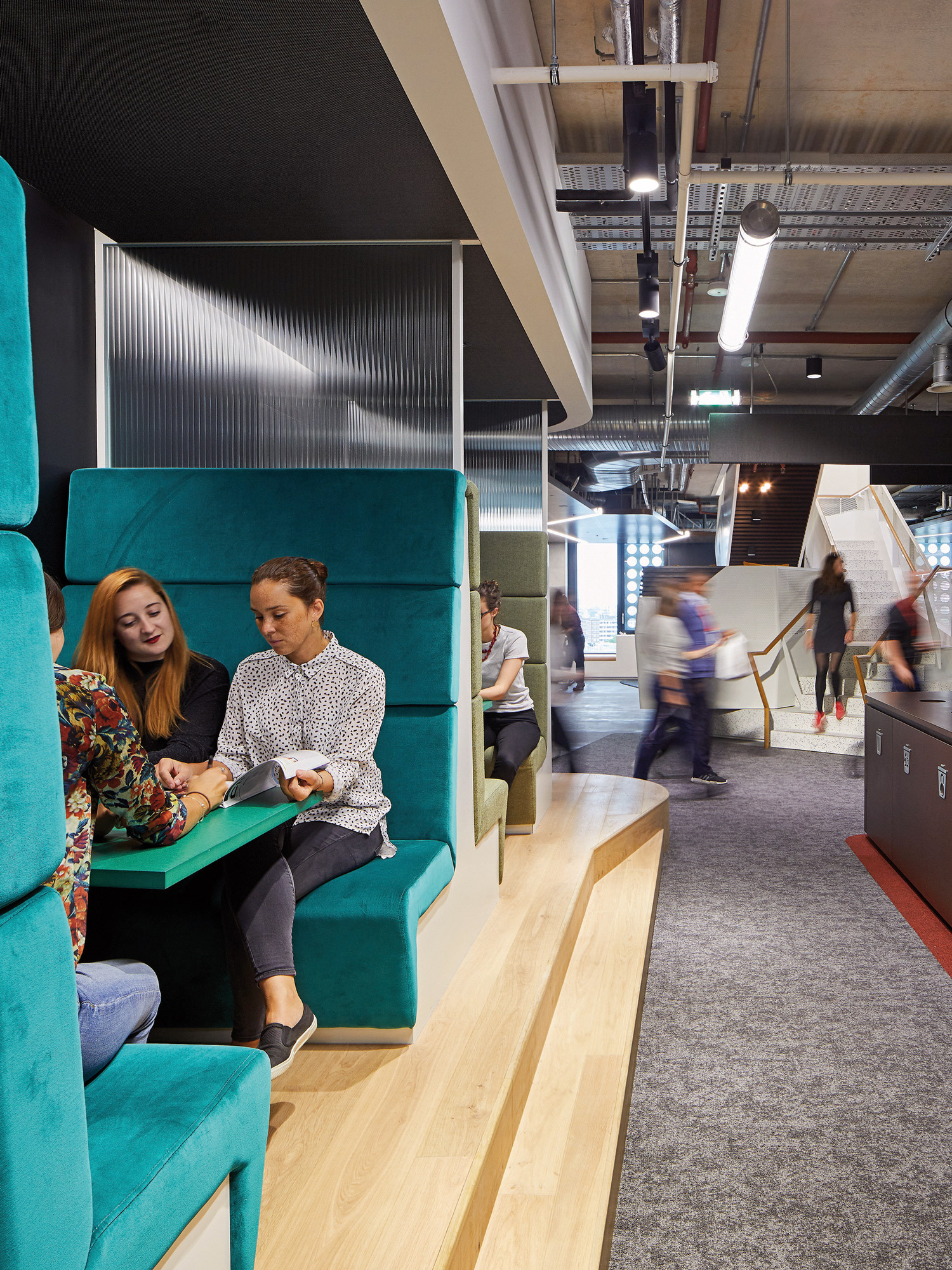 A high-backed, teal booth seating with integrated shelving offers a private workspace in an open-plan office environment, as two professional women engage in a collaborative session against a backdrop of active colleagues in motion.