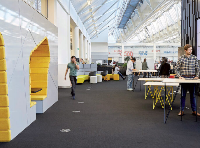 Modern office interior with an open floor plan, featuring angular lighting fixtures overhead and a vibrant yellow seating nook. Collaborative spaces with high-top tables encourage interaction, while the far wall showcases project displays under a naturally lit atrium.